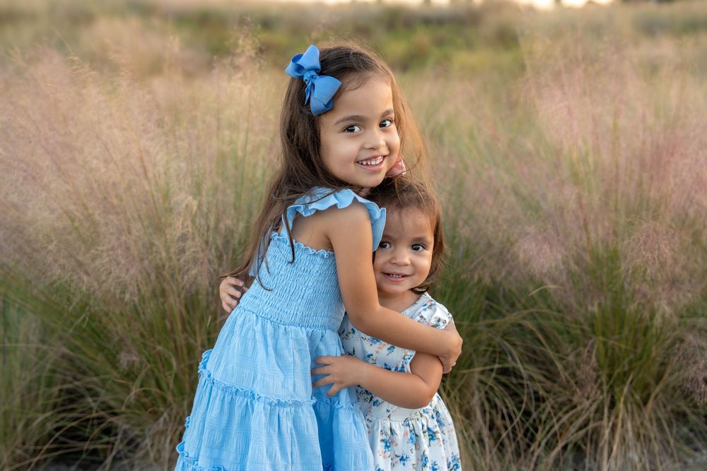 Two young sisters in blue dresses share a warm hug in a scenic outdoor field with tall pink grasses.