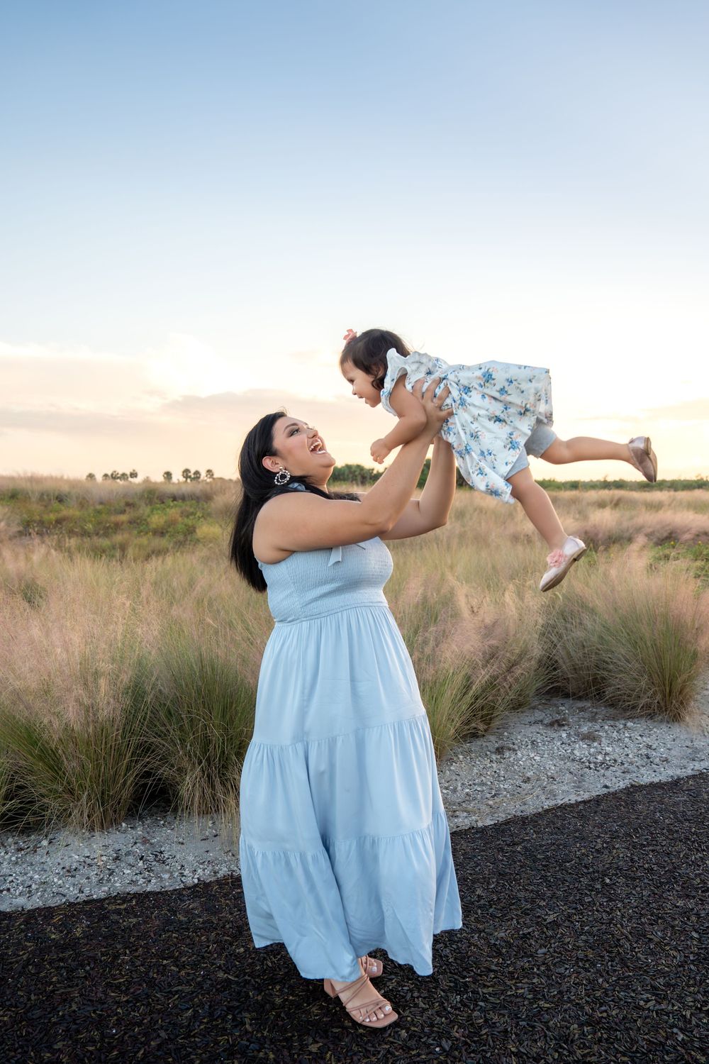 A mother in a light blue maxi dress joyfully lifts her baby girl in the air at sunset outdoors.