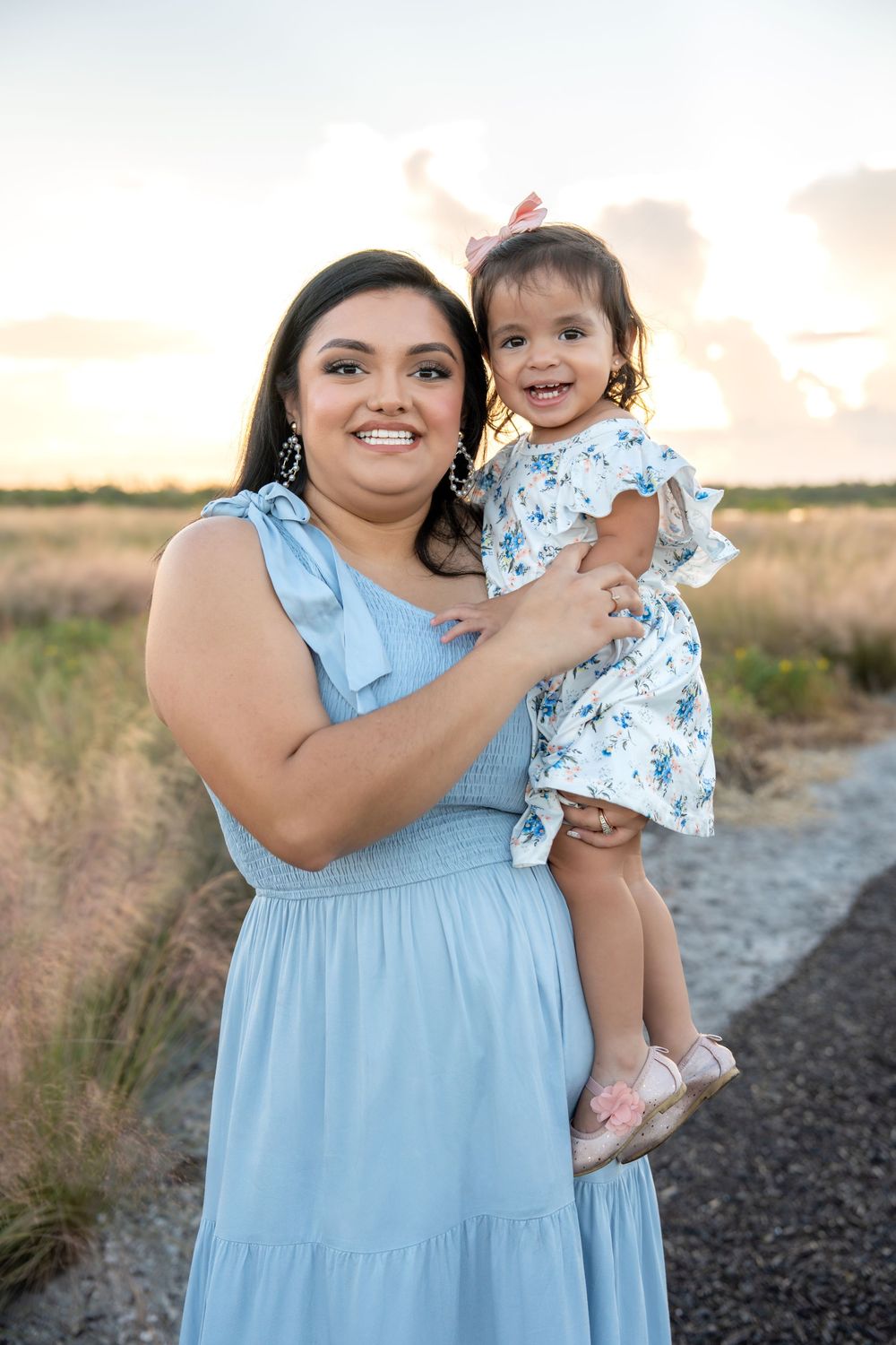 A smiling woman in a light blue dress holds a toddler girl in a floral dress at sunset in a field.