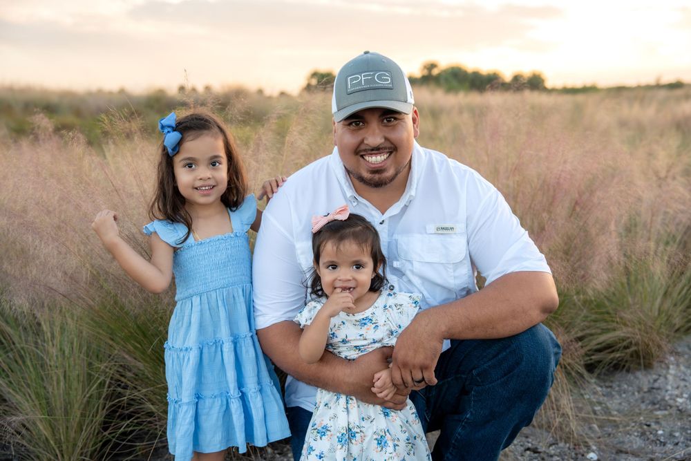A father crouches in a field at sunset, smiling with two young daughters in blue dresses, wearing a PFG cap.