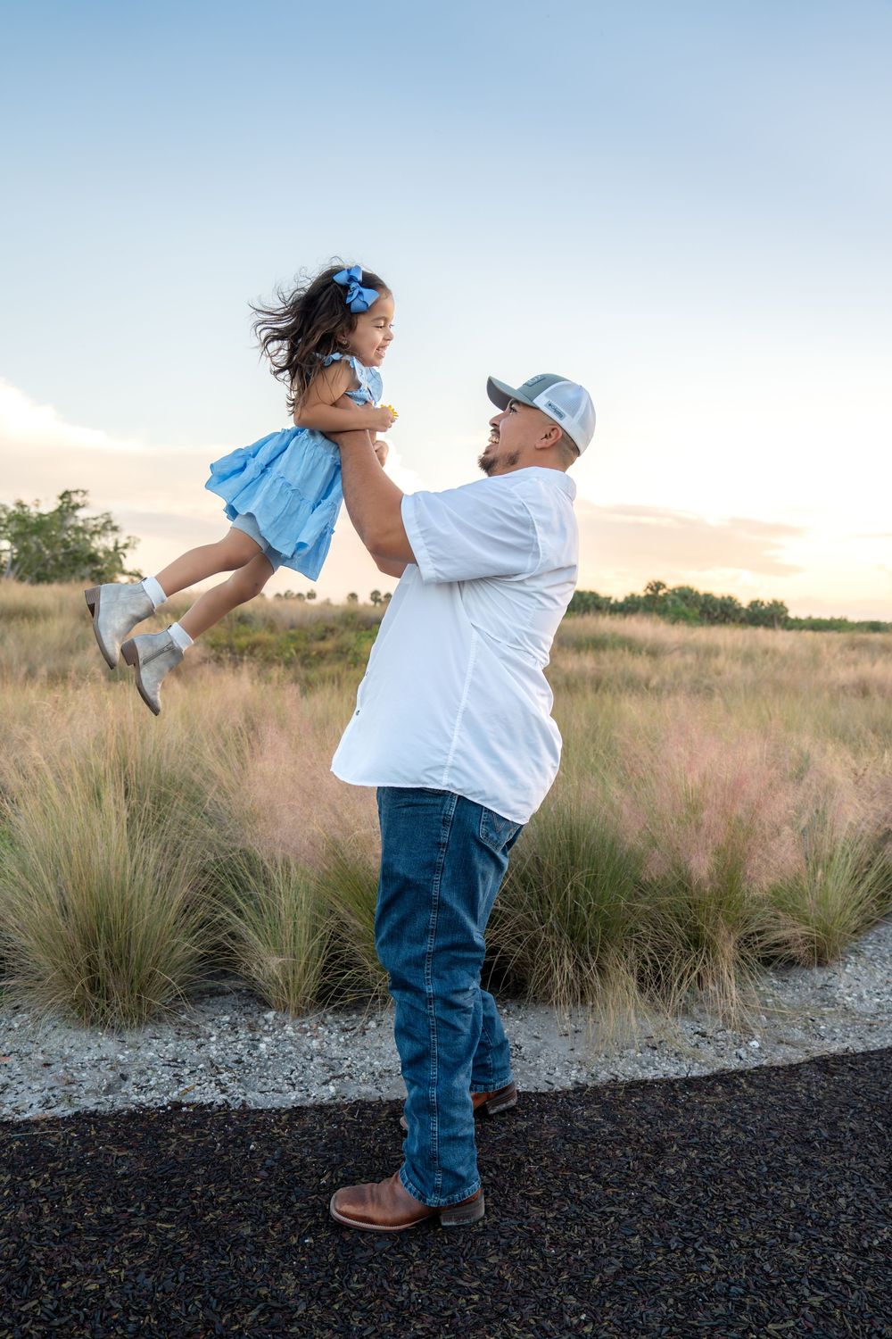 A father joyfully lifts his young daughter in a blue dress outdoors at sunset in a grassy field.