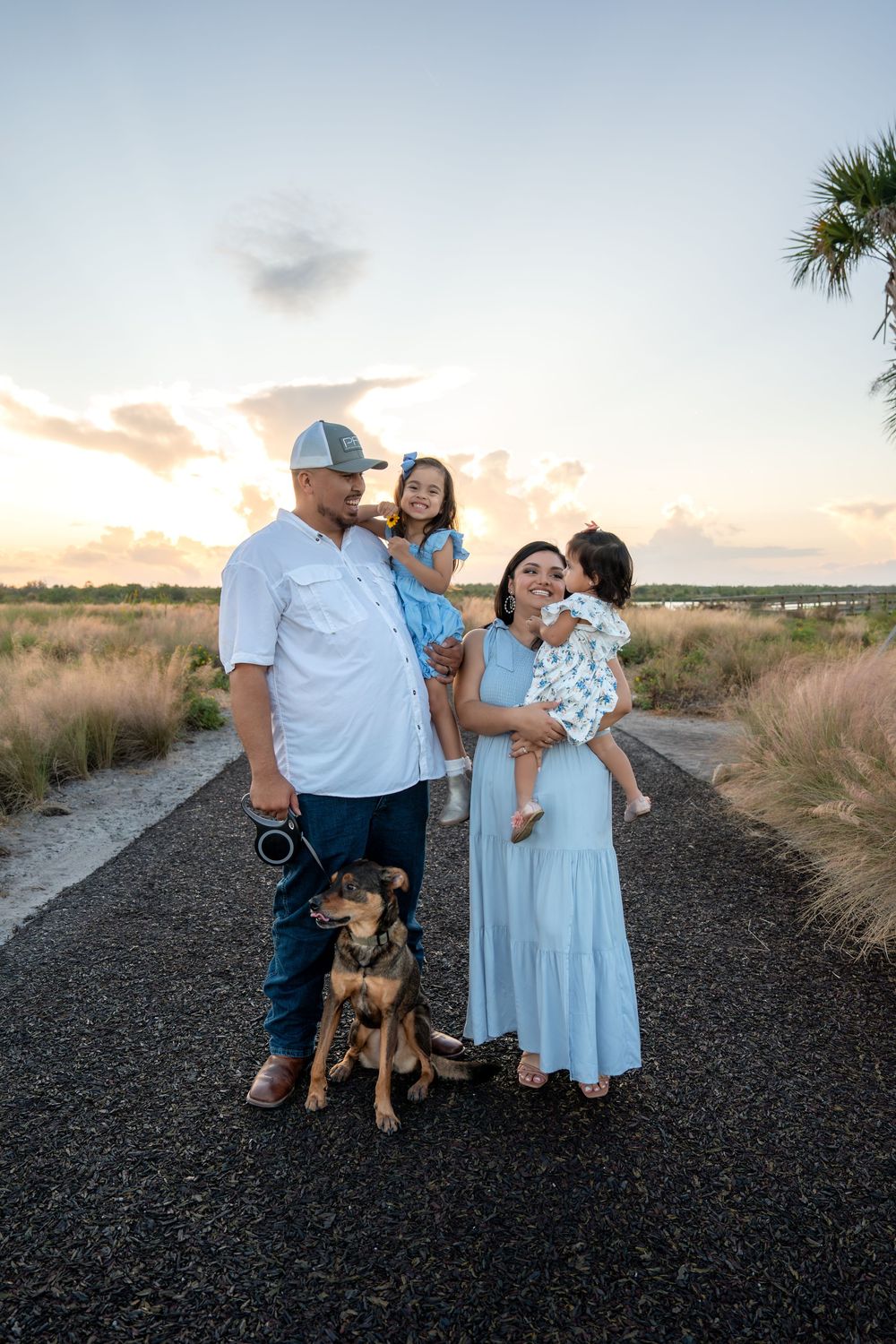 A happy family with two young daughters and a dog pose on a path at golden hour near the coast.