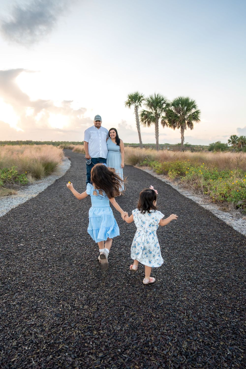 Two girls in blue dresses run toward their parents on a scenic path lined with palm trees at sunset.