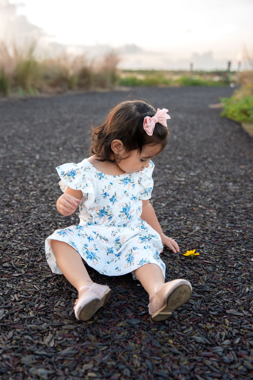 Toddler girl in floral dress and pink bow sits on gravel path outdoors.