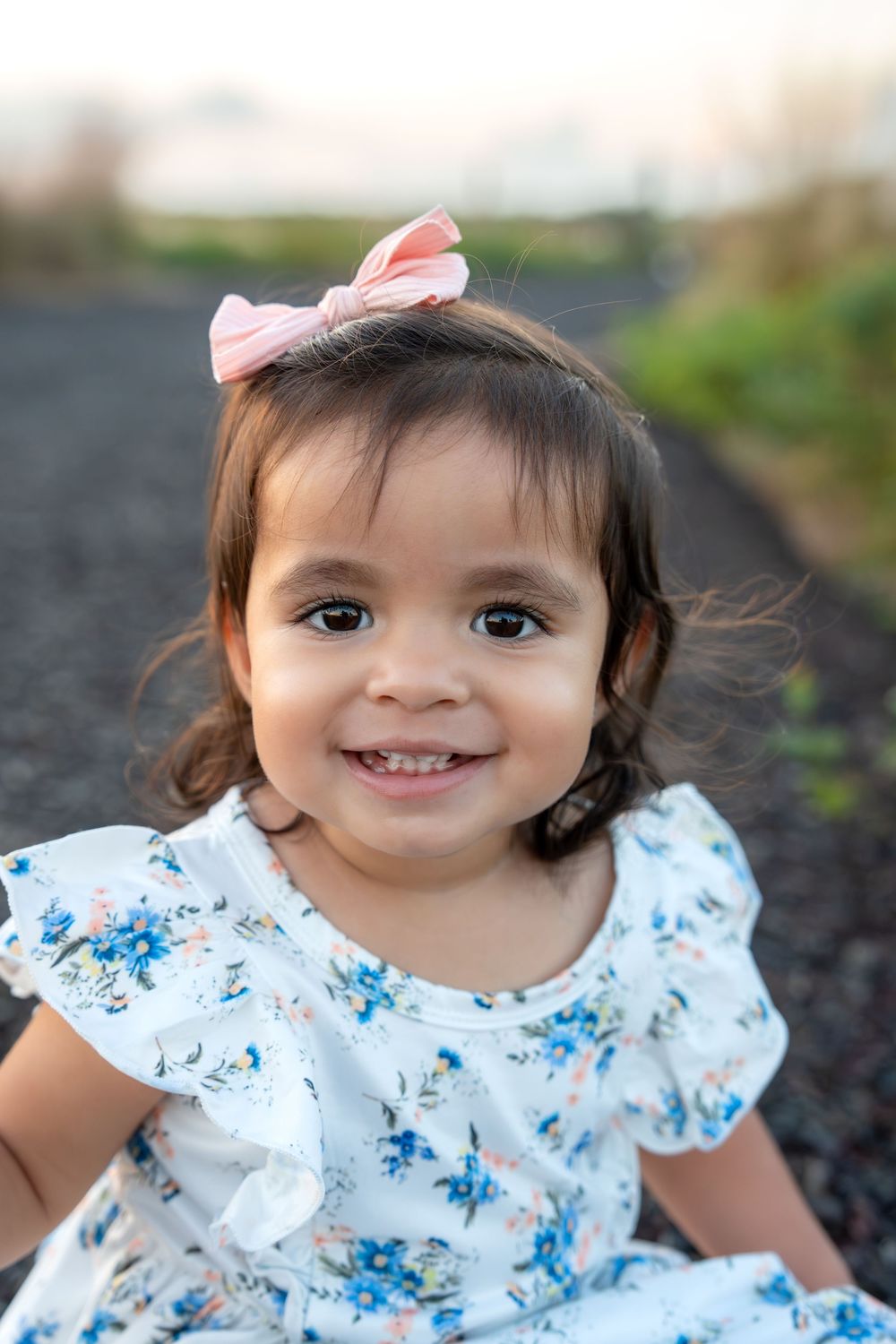 Smiling toddler girl with pink bow wearing white floral dress outdoors.