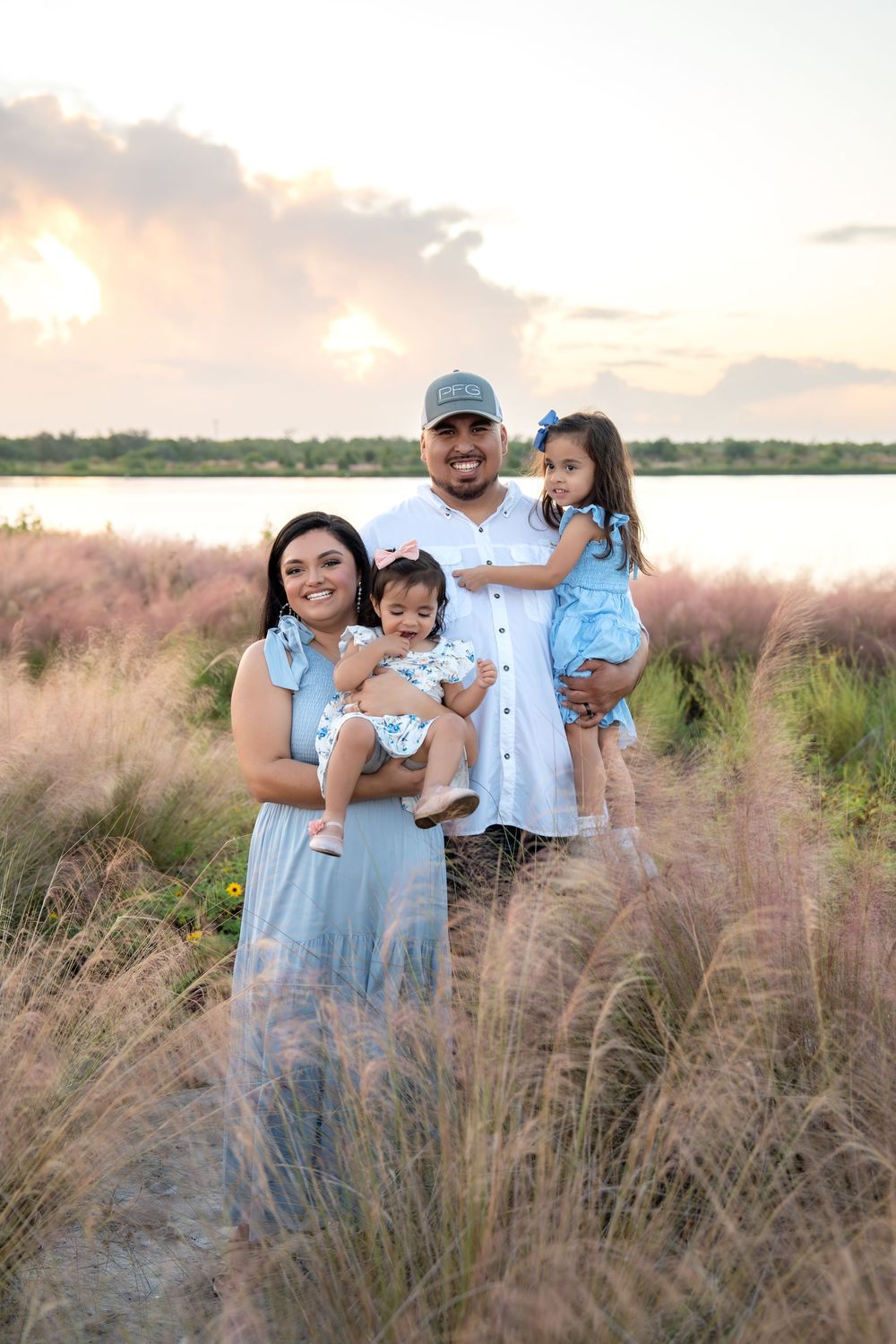Family of four poses in pink grass field at sunset, parents holding two young daughters in coordinating blue outfits.