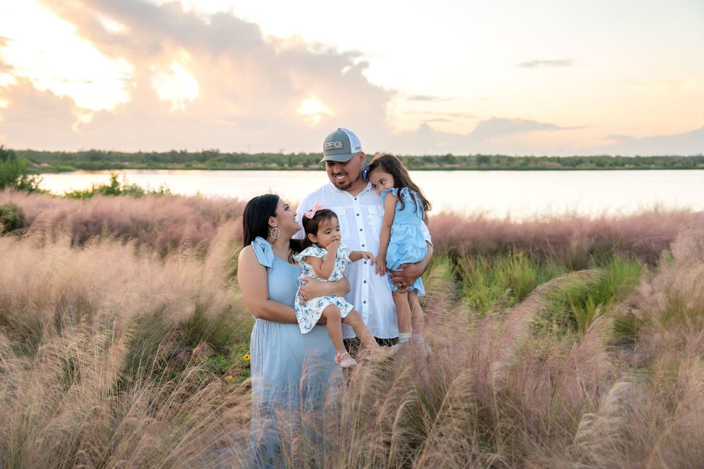 Family of four smiling together in a field of pink muhly grass at sunset near a lake.