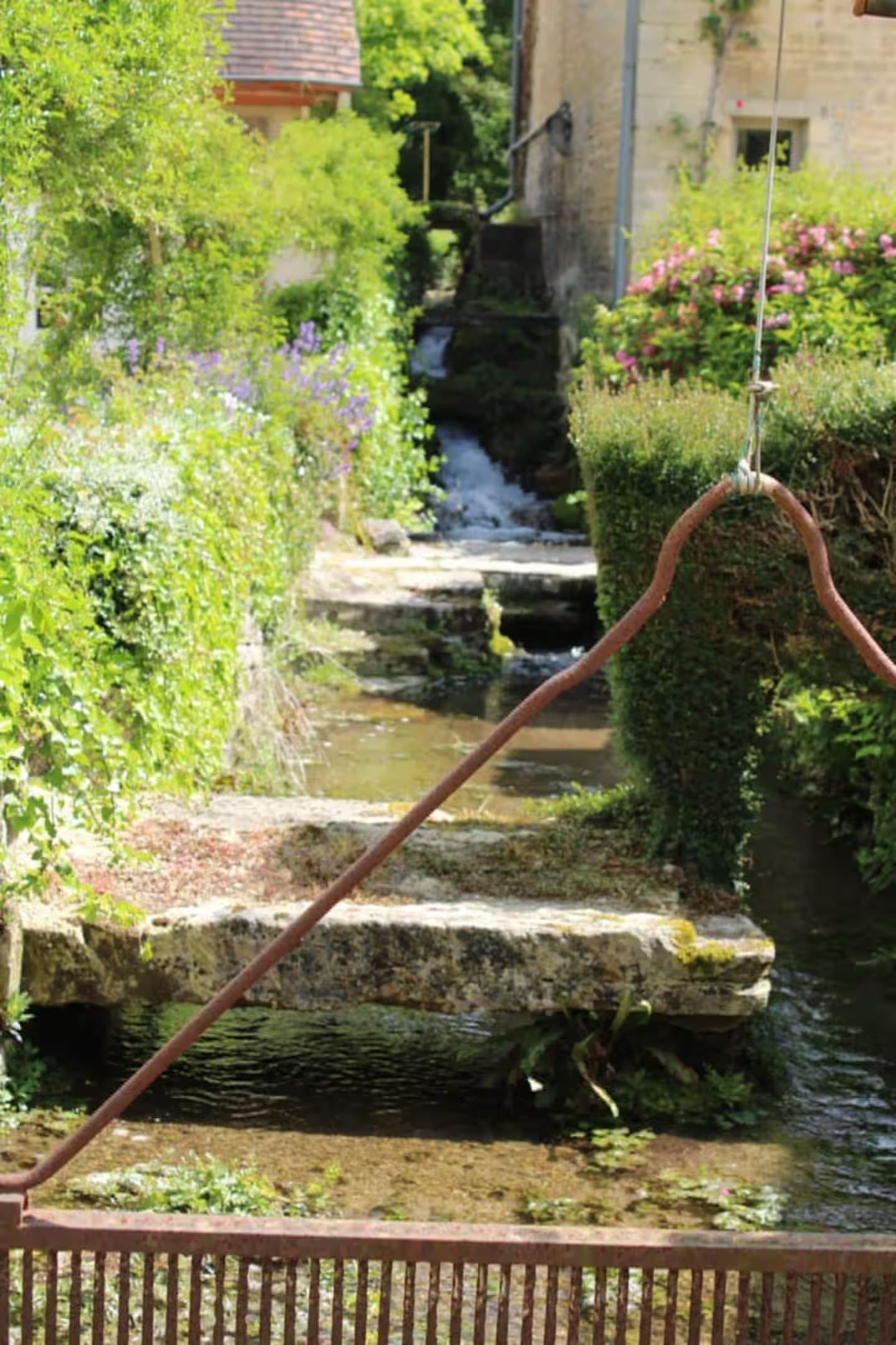 Stone steps descend beside a lush garden stream with ivy-covered walls and colorful flowers in a scenic countryside setting.