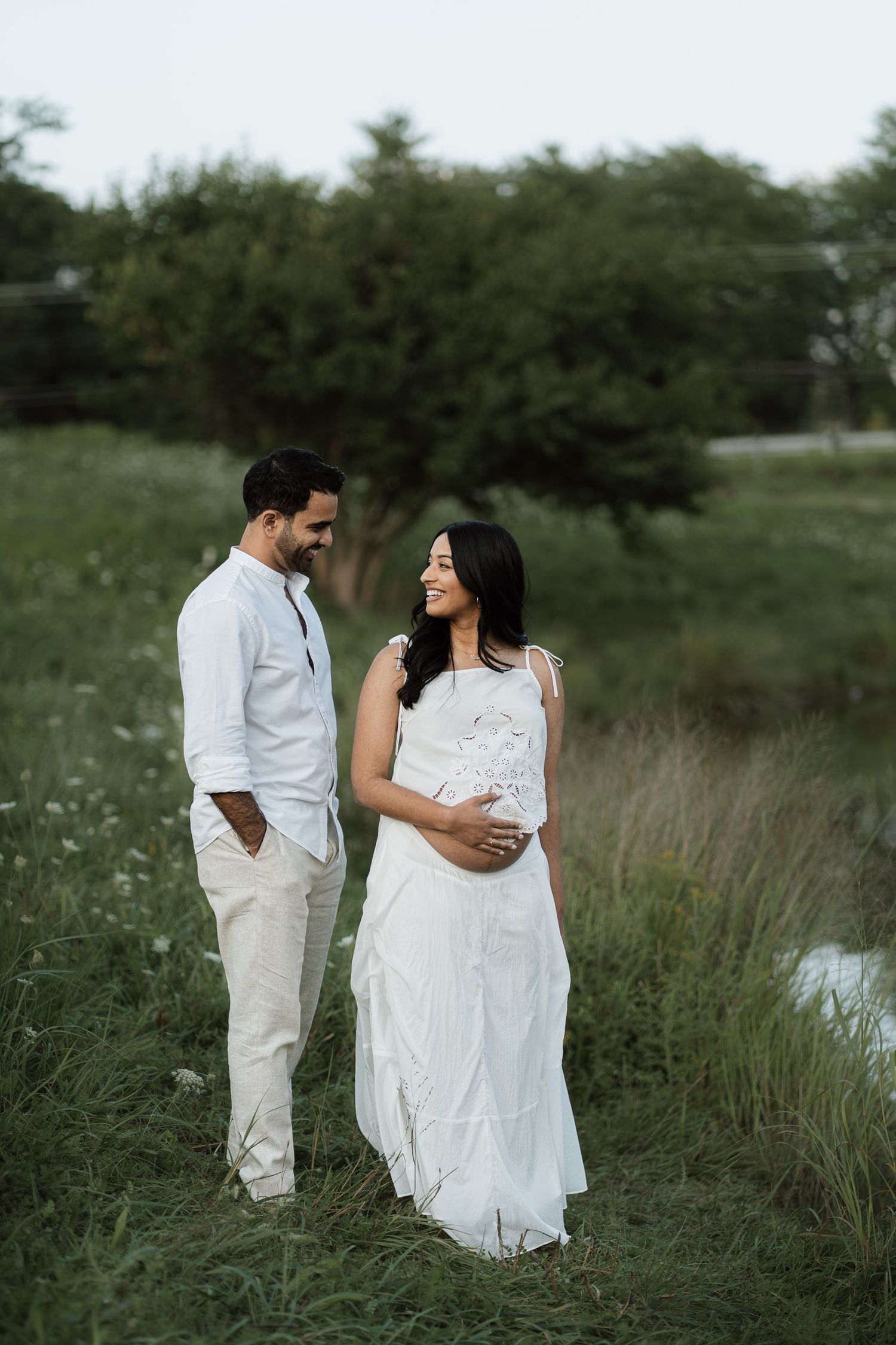 Couple in white outfits standing in a green field, pregnant woman cradling her baby bump during a maternity photoshoot.
