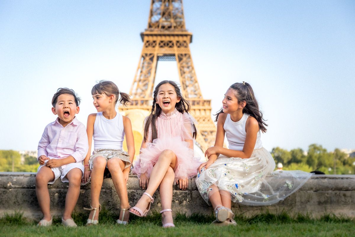 Four joyful Asian children laughing and sitting together on a stone ledge with the Eiffel Tower in Paris behind them.