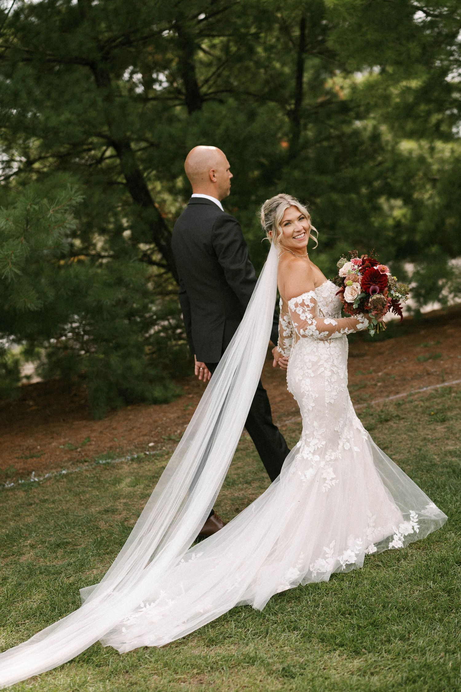 Bride in lace mermaid gown with long veil holds bouquet, groom in tuxedo, outdoor wedding portrait.
