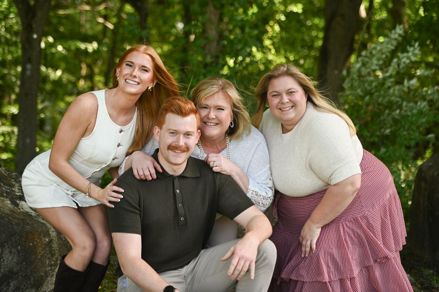A joyful family of four poses outdoors among lush green trees, smiling warmly for a portrait photo.
