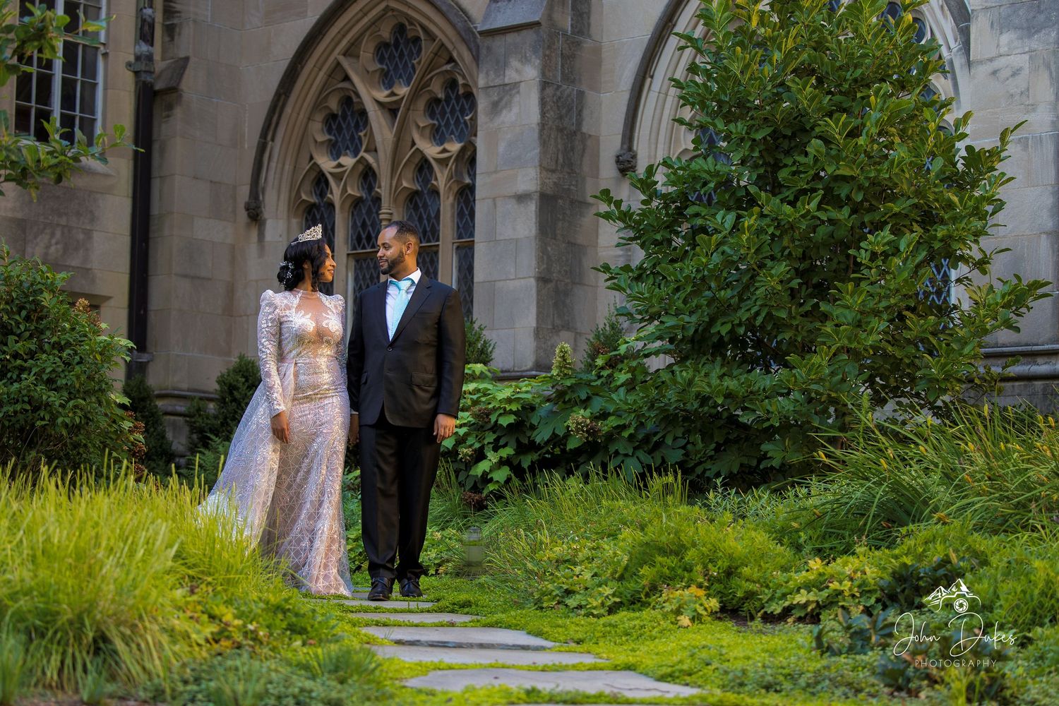 Bride and groom stand together in a lush garden beside a Gothic stone church on their wedding day.