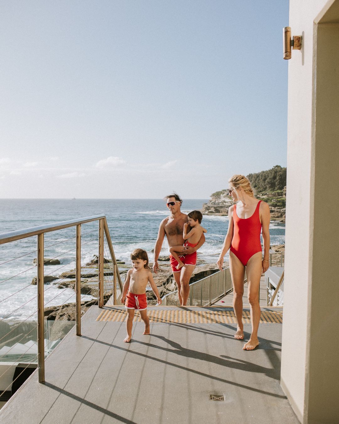 Family in matching red swimwear on oceanfront deck with rocky coastline and blue sea in background.