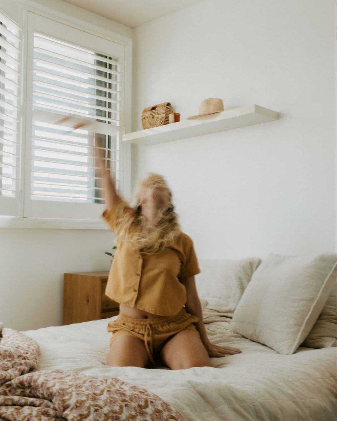 A blonde woman in a mustard outfit joyfully tosses her hair while kneeling on a white bed in a bright bedroom.