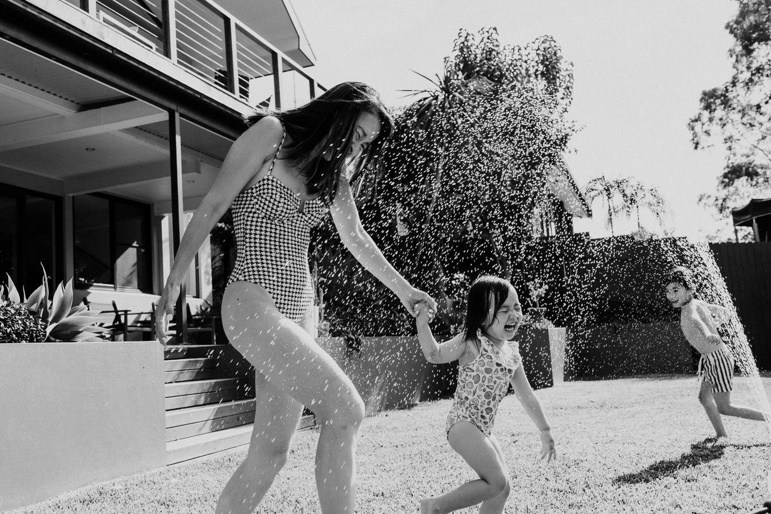Black and white photo of women and children playing in sprinkler water in backyard on sunny day.