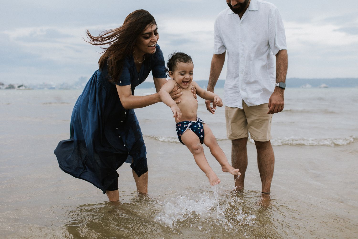 Family playing at beach, parents swinging baby over ocean waves, woman in blue dress, man in white shirt.