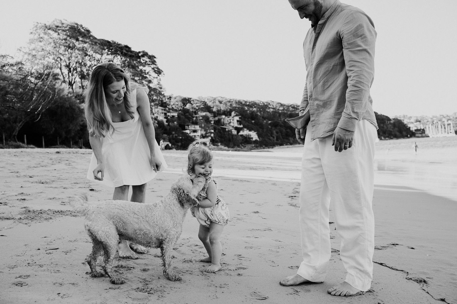 Family with toddler and dog enjoying a playful moment on a sandy beach in black and white photo.
