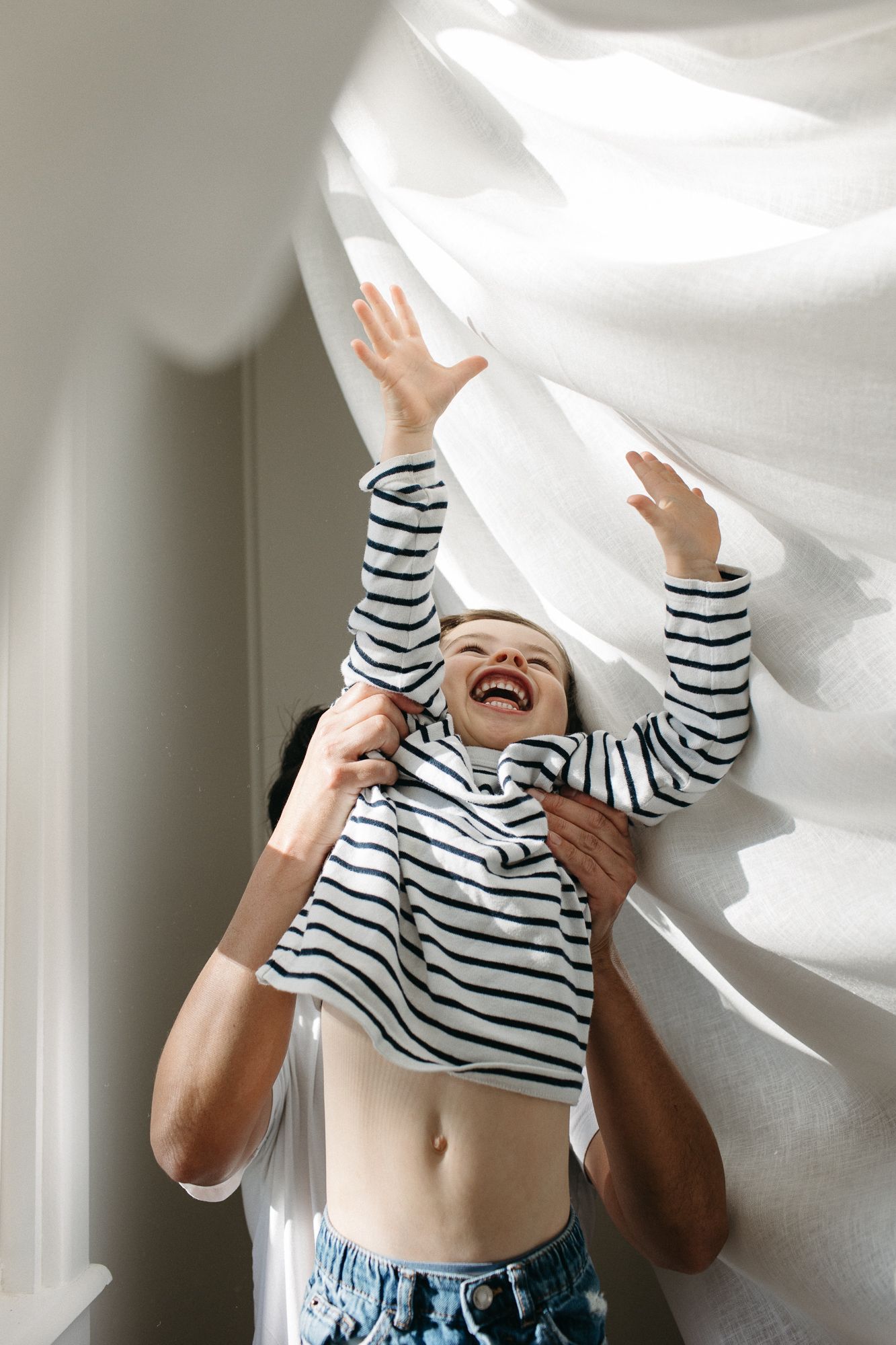 Joyful woman in striped top laughing with arms raised near billowing white curtains in bright room.