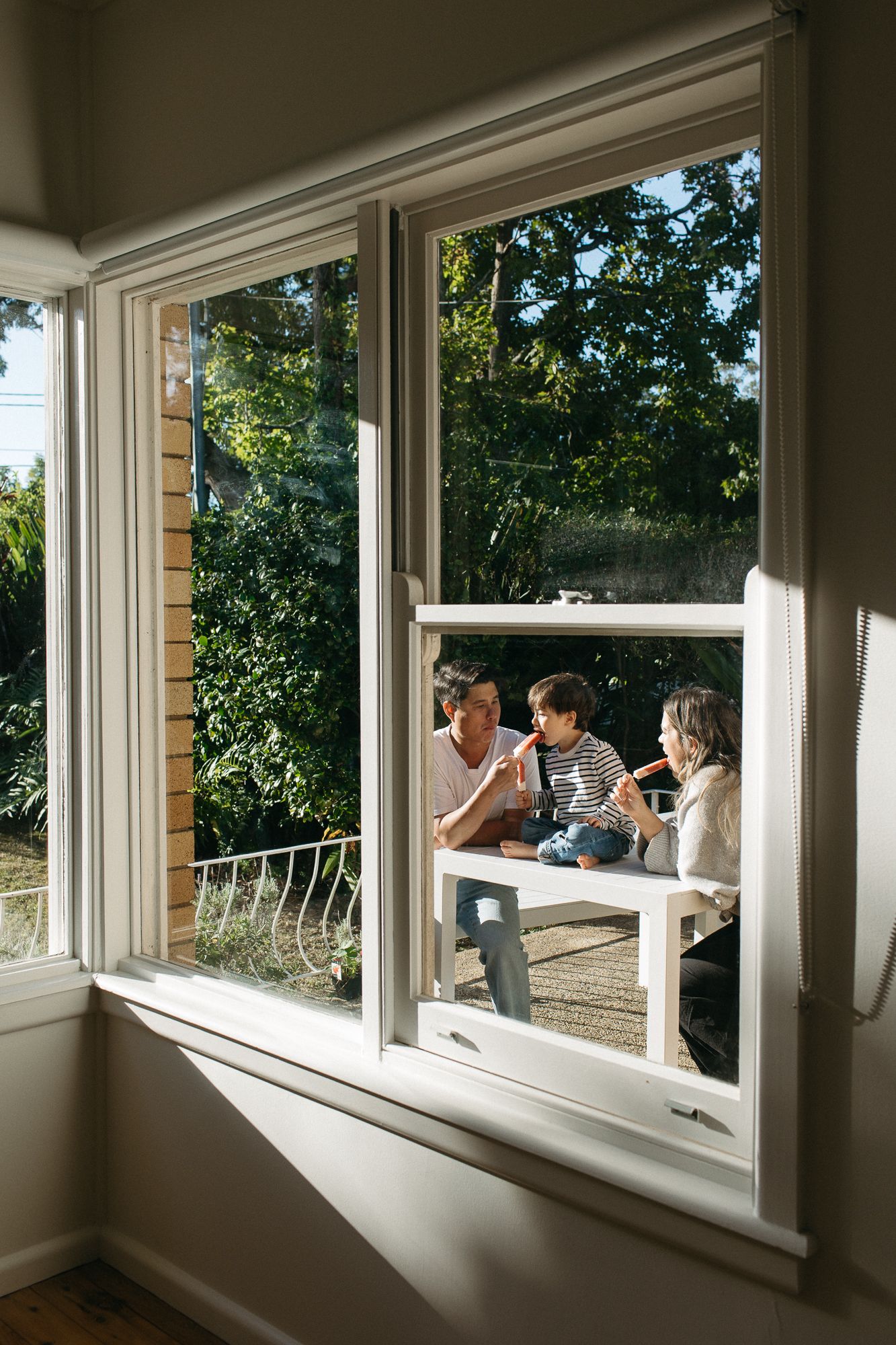 Family of three enjoying popsicles at outdoor table, viewed through sunny window with lush garden backdrop.