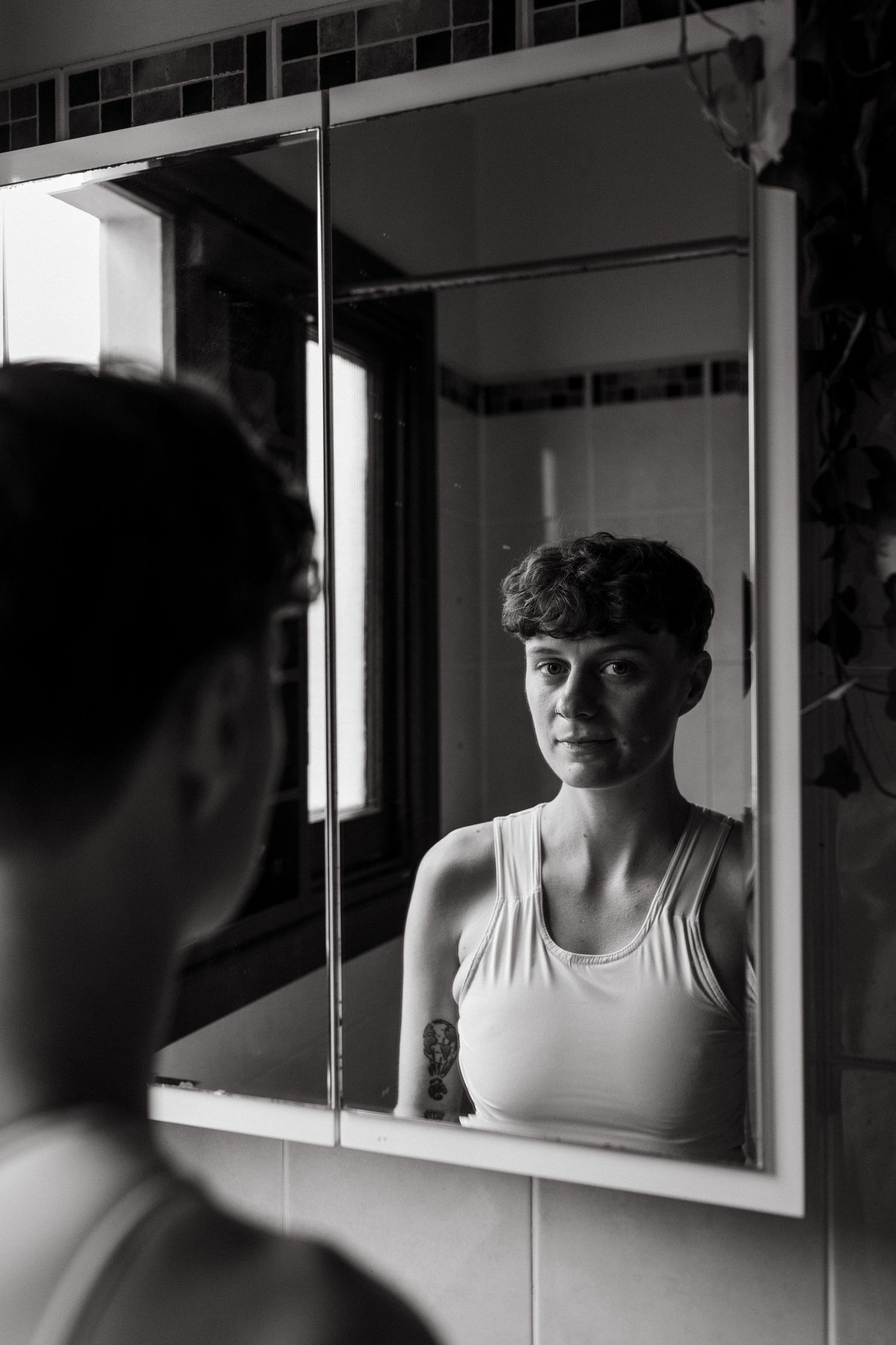 Black and white photo of a person with short hair reflecting in a bathroom mirror, wearing a white tank top.