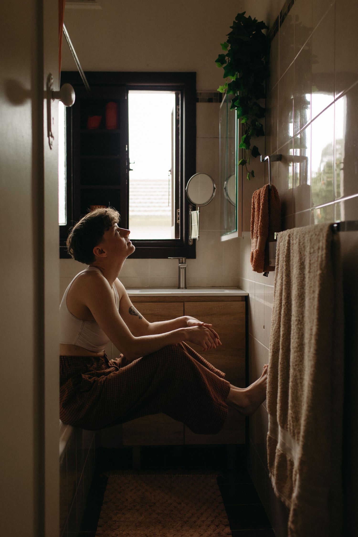 Person sitting on bathroom counter in natural light, wearing white tank top, surrounded by plants and towels.