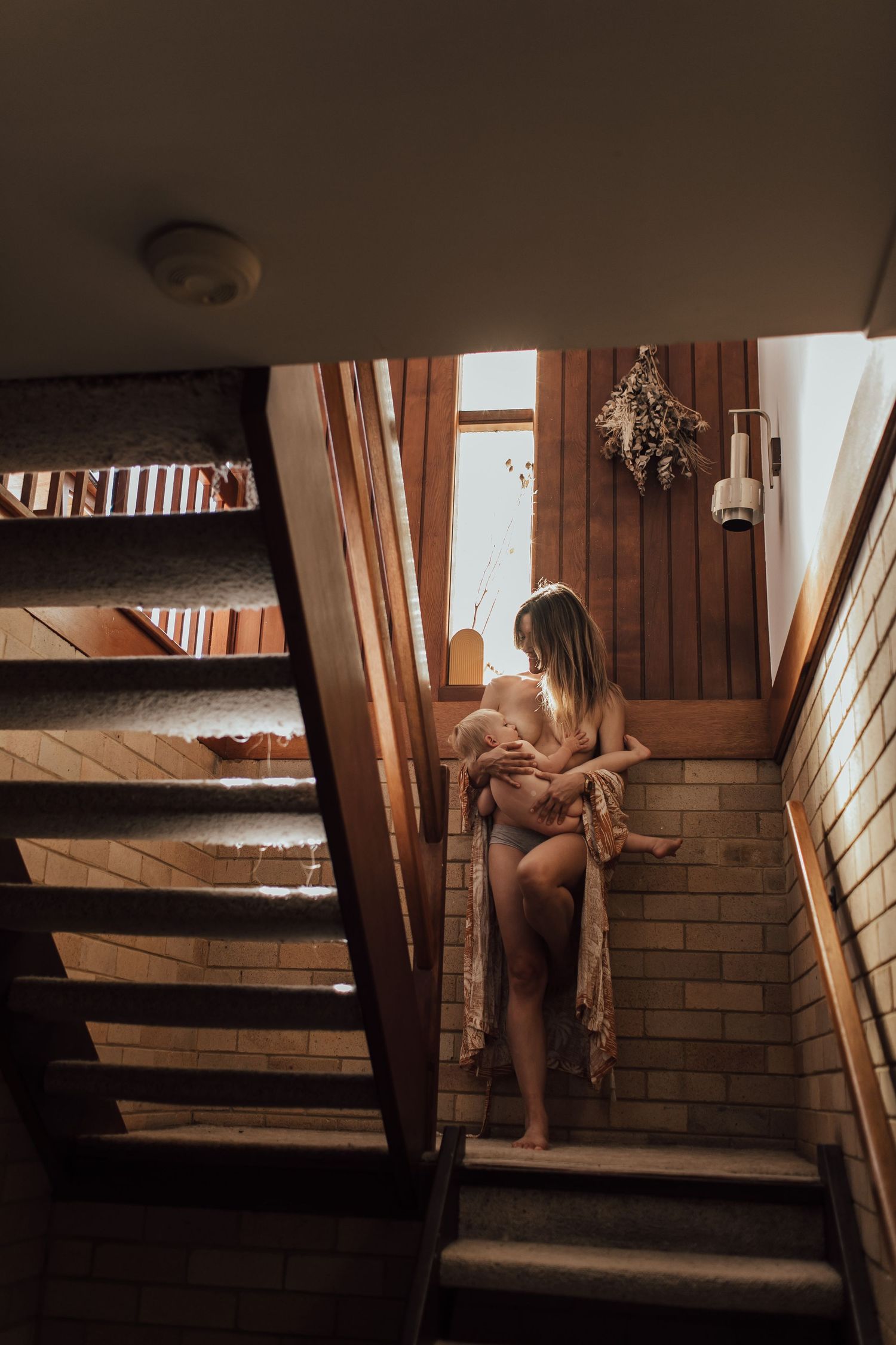 A woman sits on stairs holding a baby in a warm, rustic wooden interior with soft natural light.