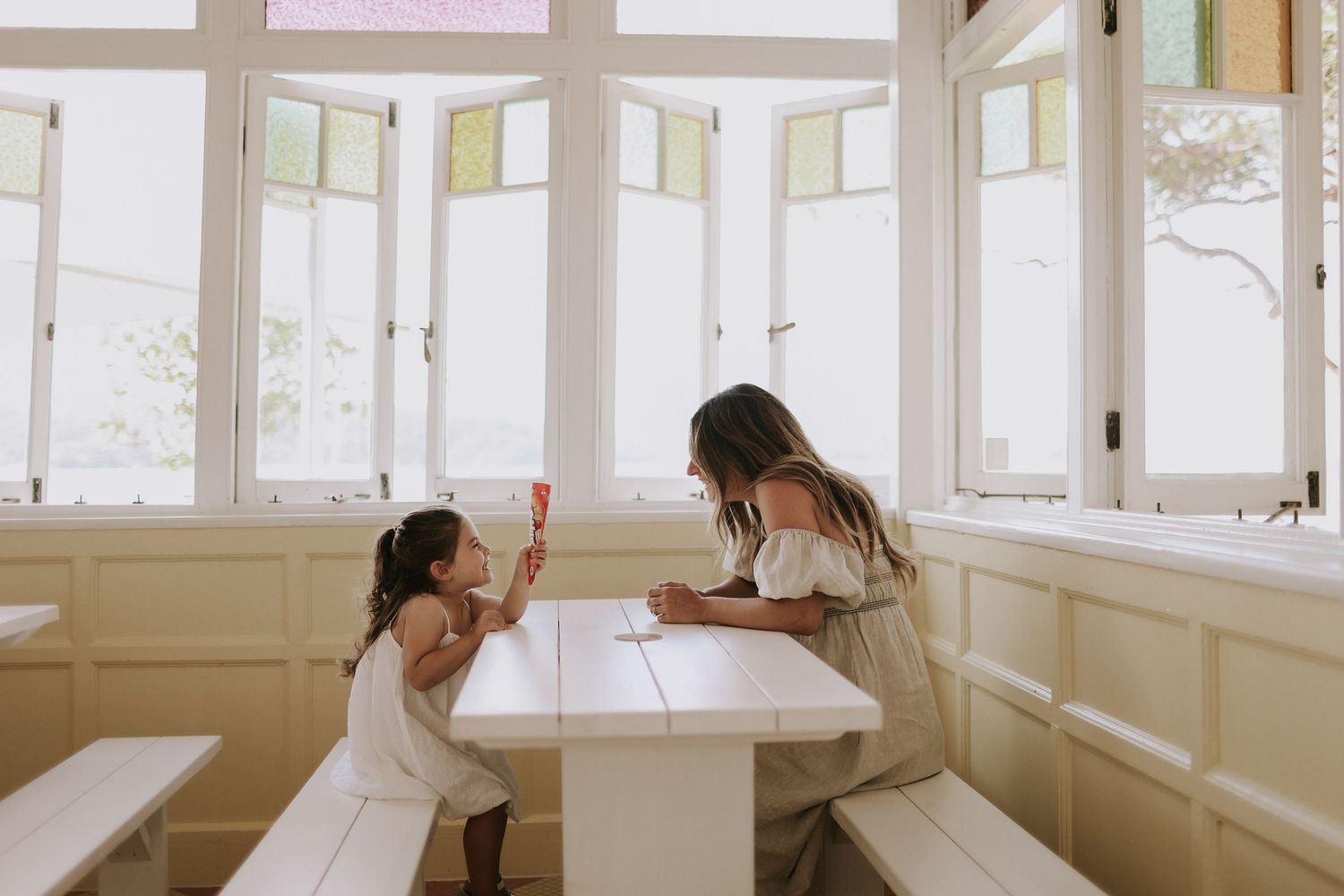 Woman and young girl sharing ice cream at a white table by bright windows in a cozy cafe.