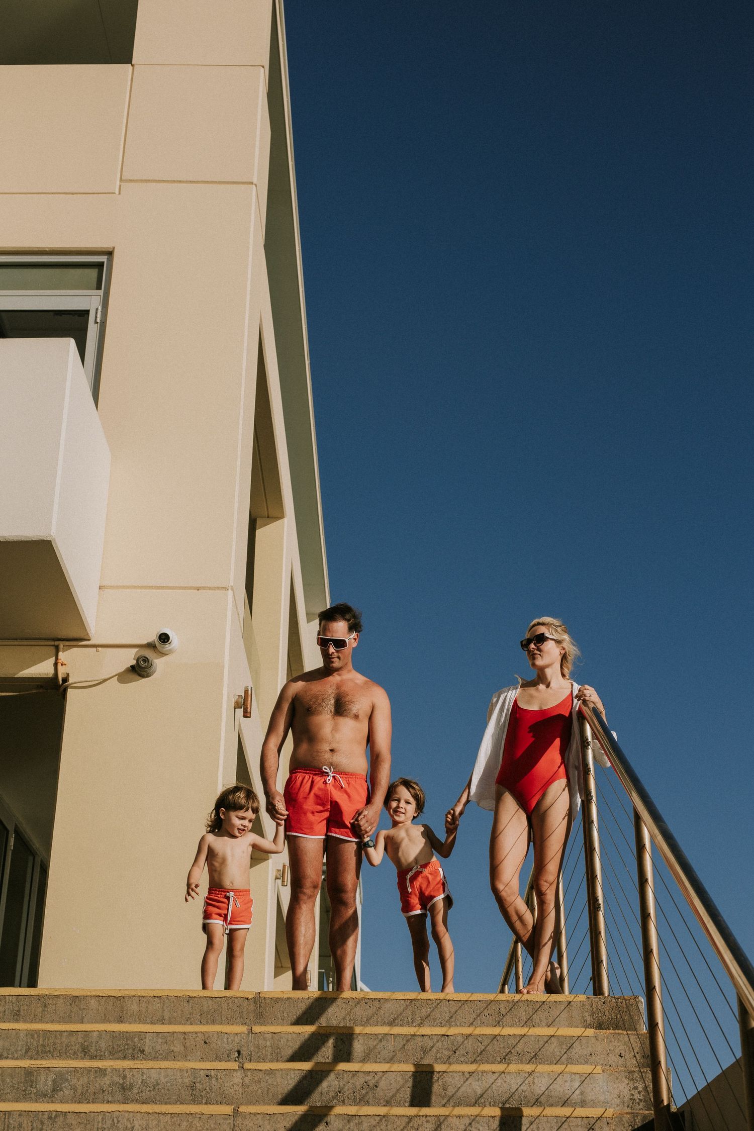 Family in red swimwear descending outdoor steps of a resort building under a clear blue sky.