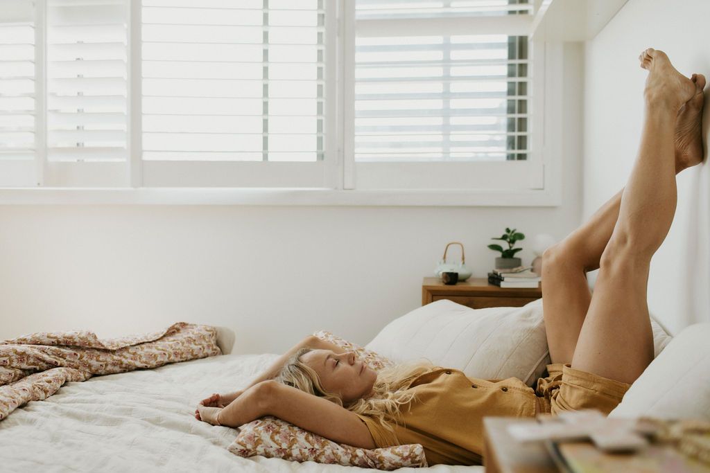 Woman relaxing on a white bed with legs raised, wearing a yellow outfit in a bright, airy bedroom with shuttered windows.