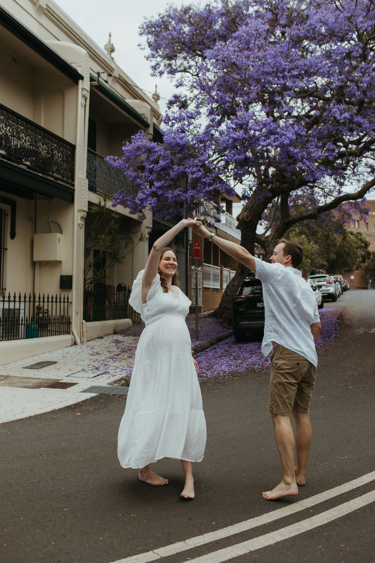 Couple dancing barefoot in street under blooming purple jacaranda tree, woman in white dress, Sydney.