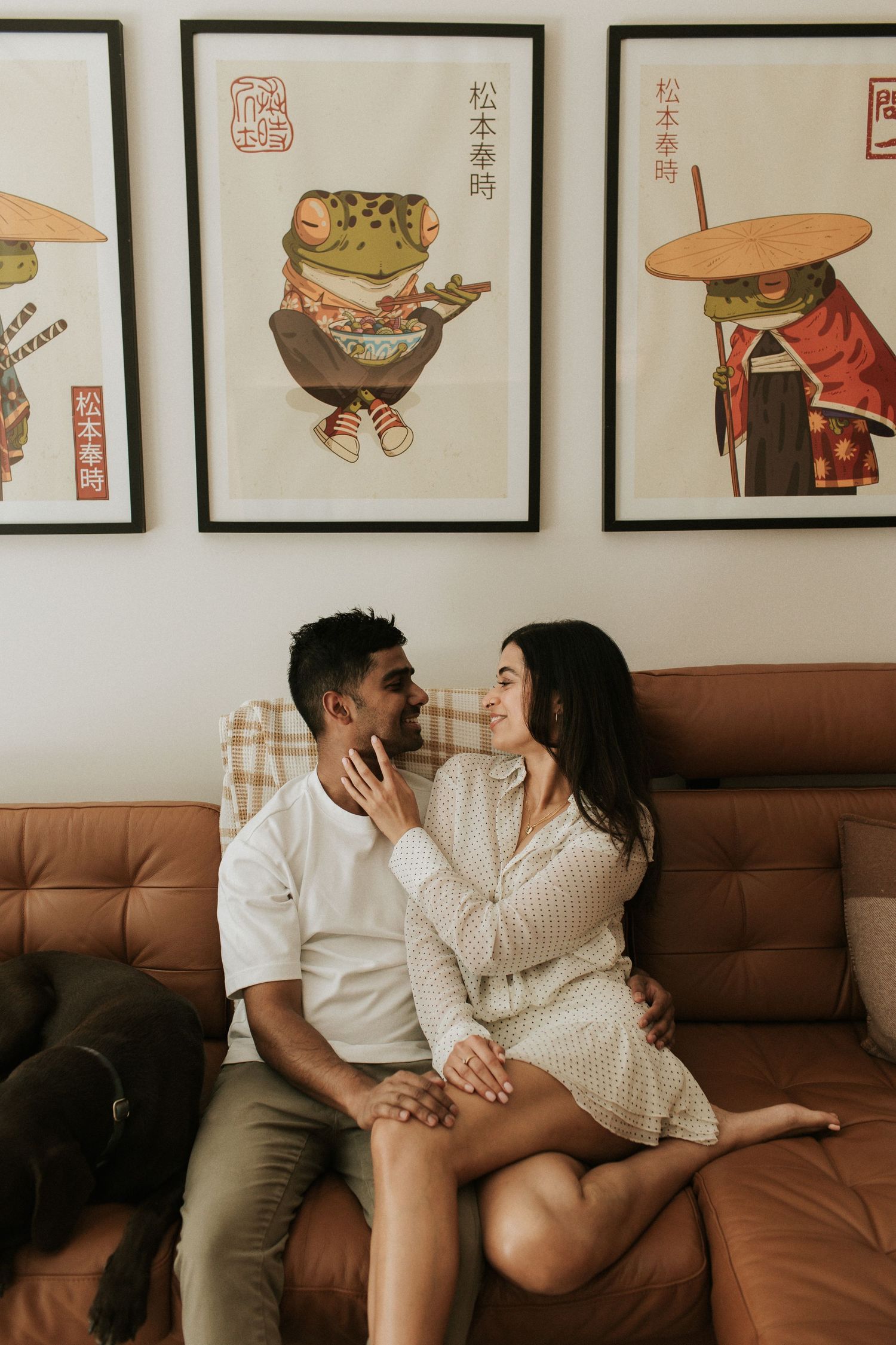 Couple sitting close on a brown leather sofa, smiling at each other, with Japanese frog art prints on the wall behind them.