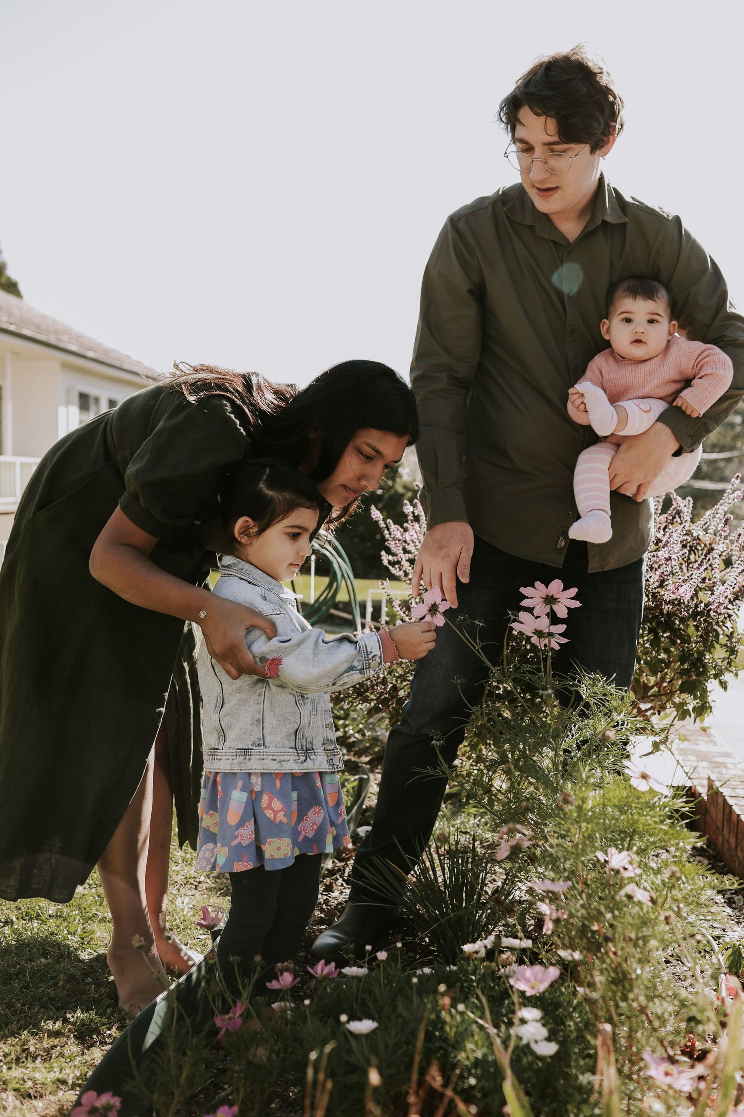 Family of four admiring pink flowers in garden, parents with toddler and baby on sunny day.