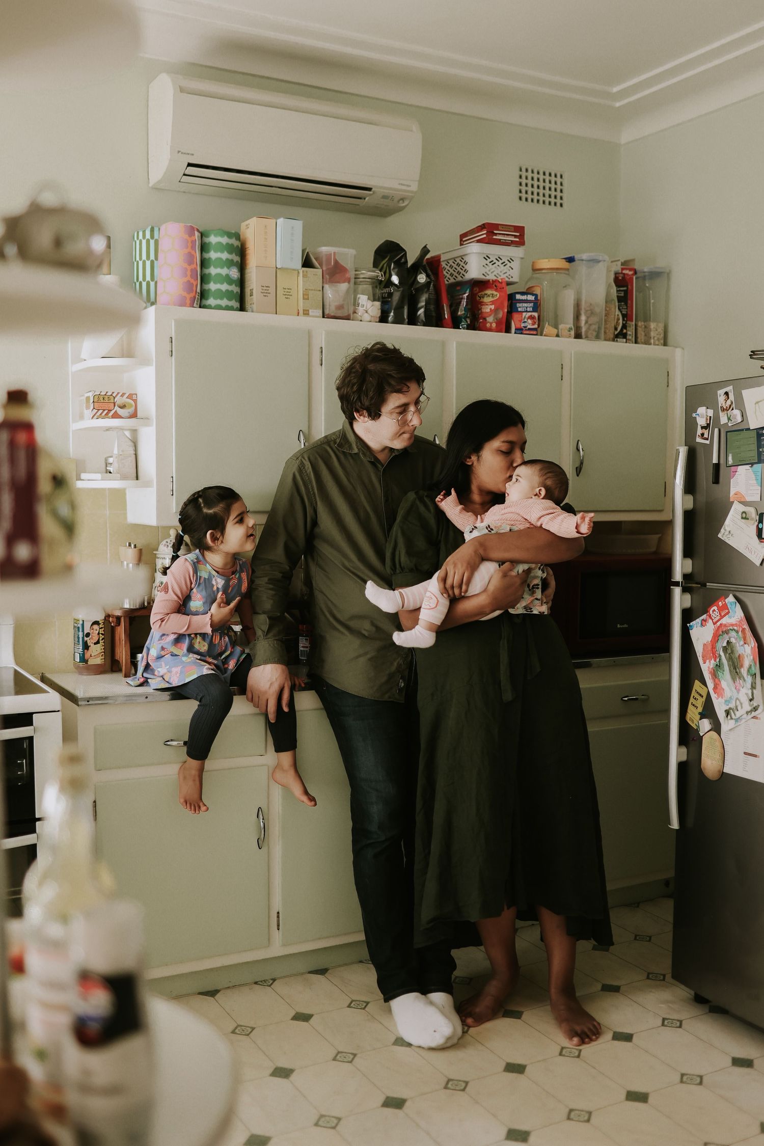 Young family with two children gathered in a cozy kitchen, parents holding a baby while older child sits on counter.