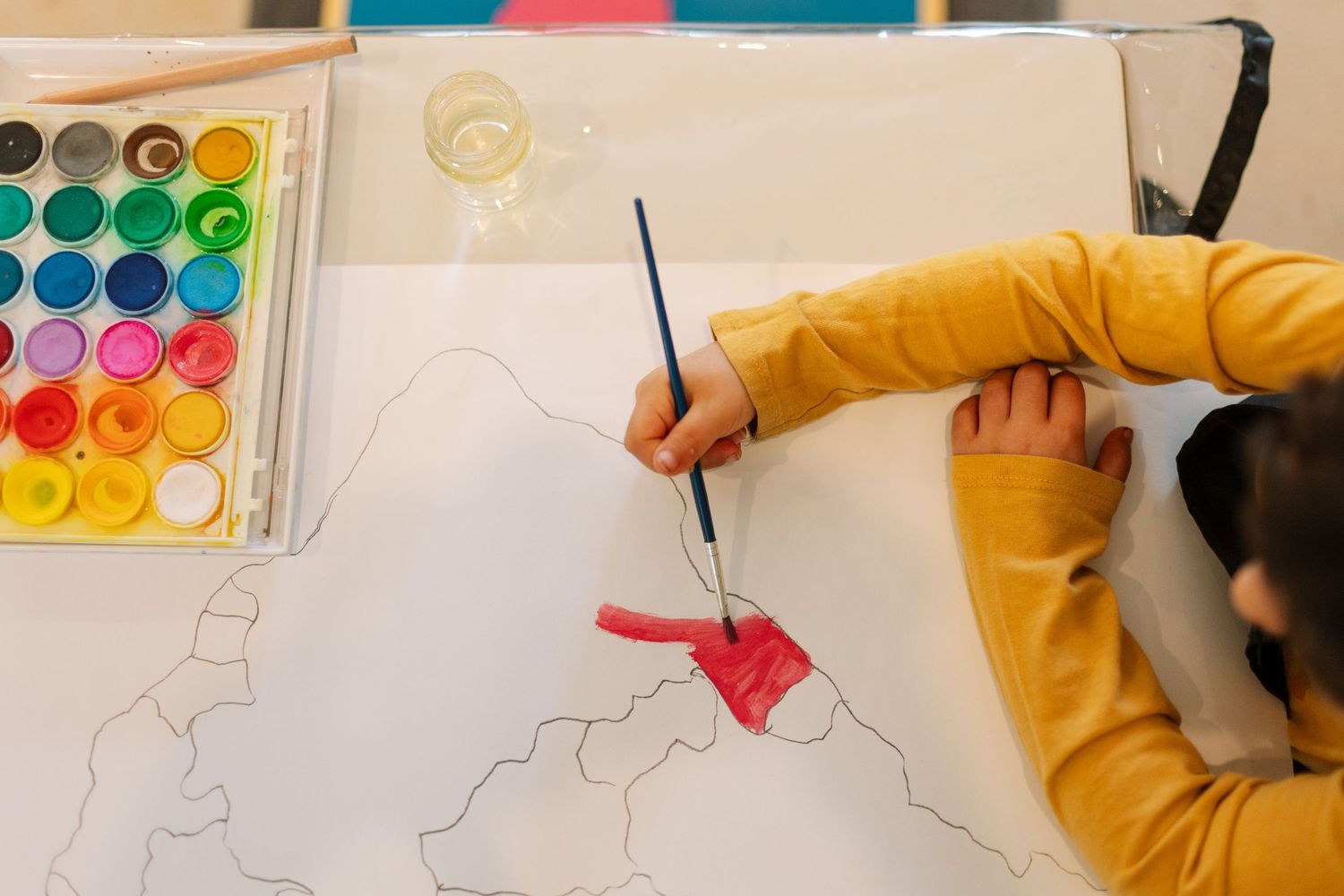 Child in yellow sweater painting a map with red watercolor paint, colorful paint palette visible on table.