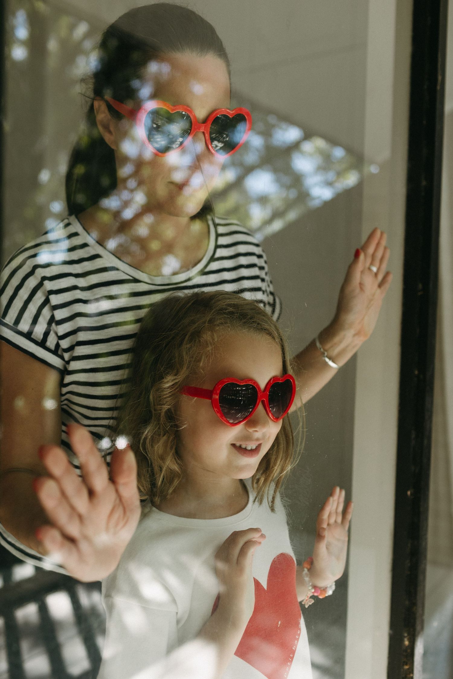 Mother and daughter wearing matching red heart-shaped sunglasses, smiling and pressing hands against a glass window.