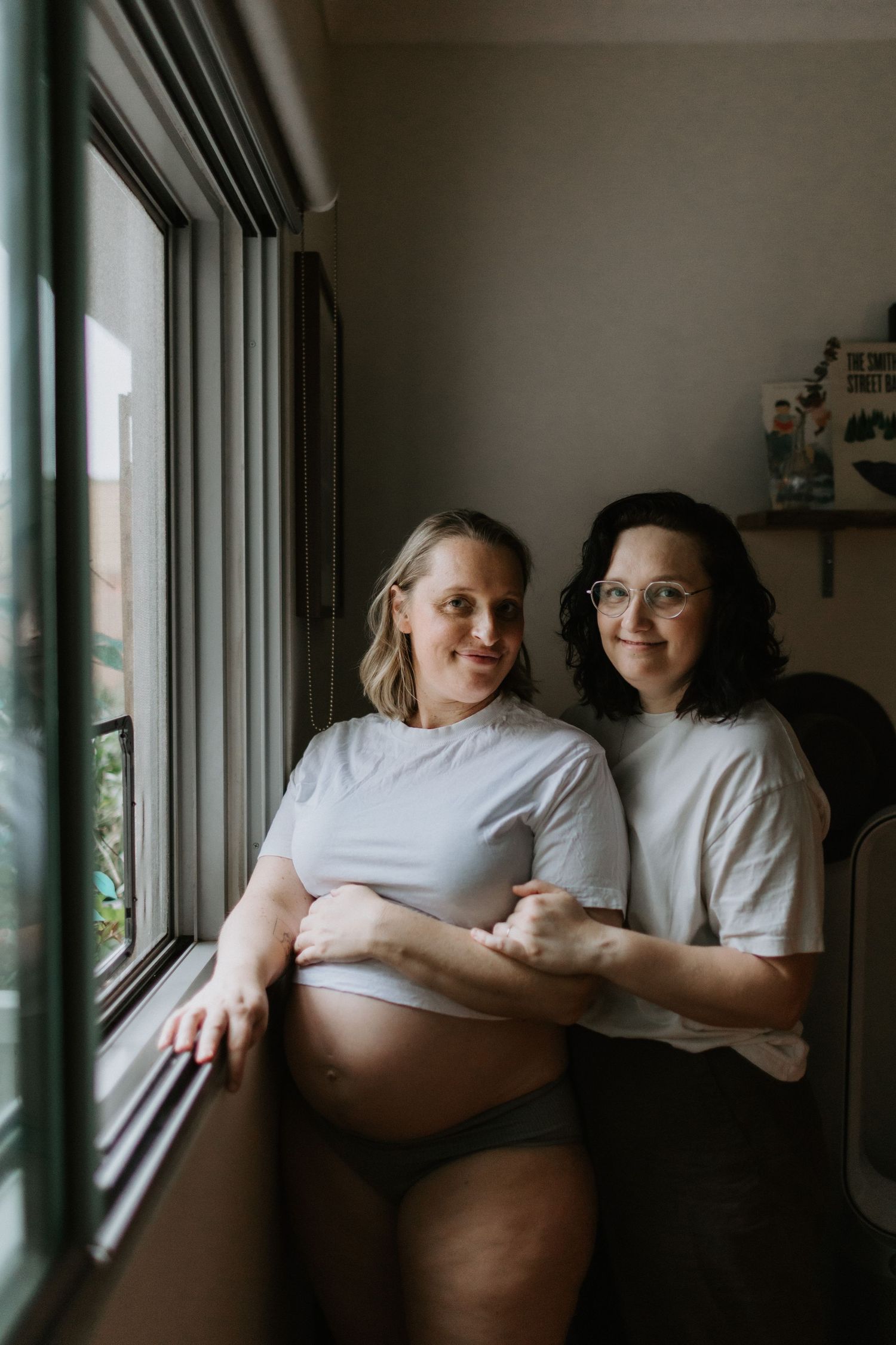 Two women pose by a window, one pregnant in a white tee, the other embracing her from behind, both smiling warmly.