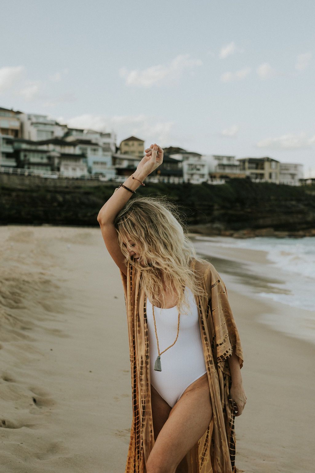 Woman in white swimsuit and boho kimono posing on sandy beach with coastal buildings in background.