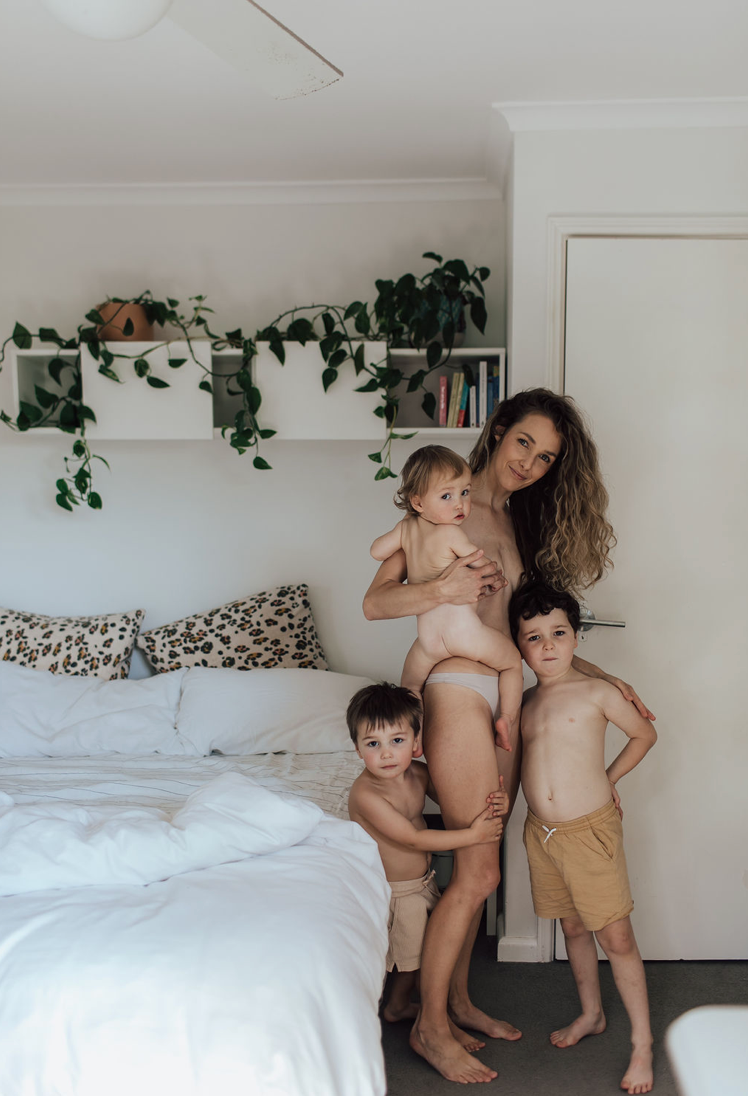 A mother holds her baby while two young boys hug her in a bright white bedroom with plants.