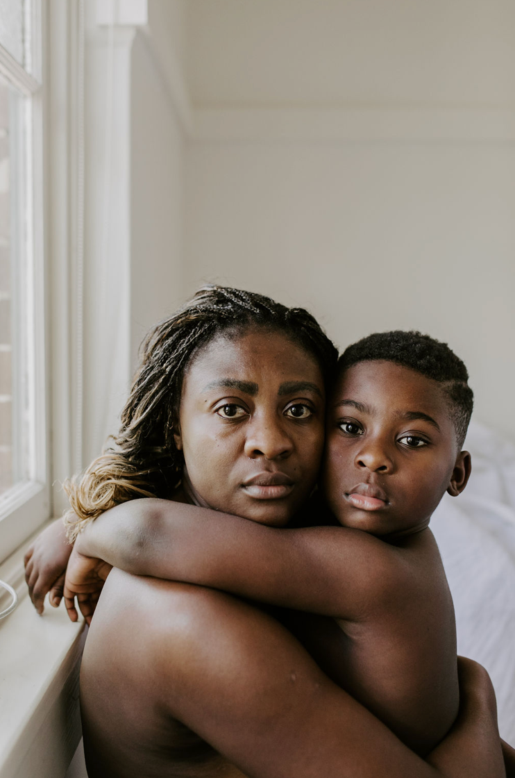 A mother and young son share a tender embrace near a bright window, both looking directly at the camera.