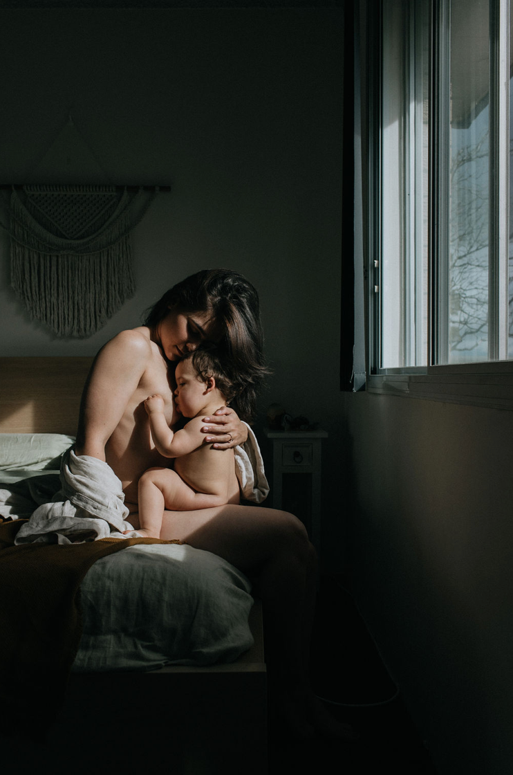 A mother tenderly holds her baby on a sunlit bed near a window, sharing an intimate bonding moment.