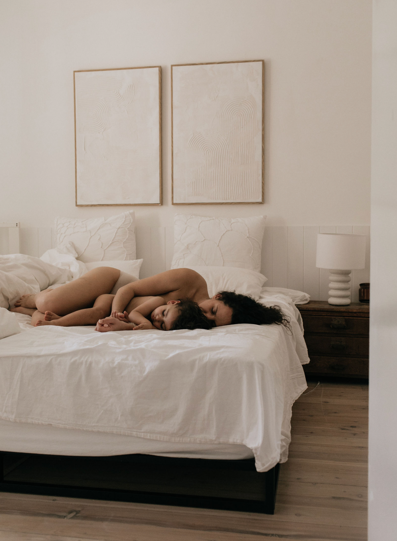 A person lying curled up on a white bed in a minimalist bedroom with framed art above.