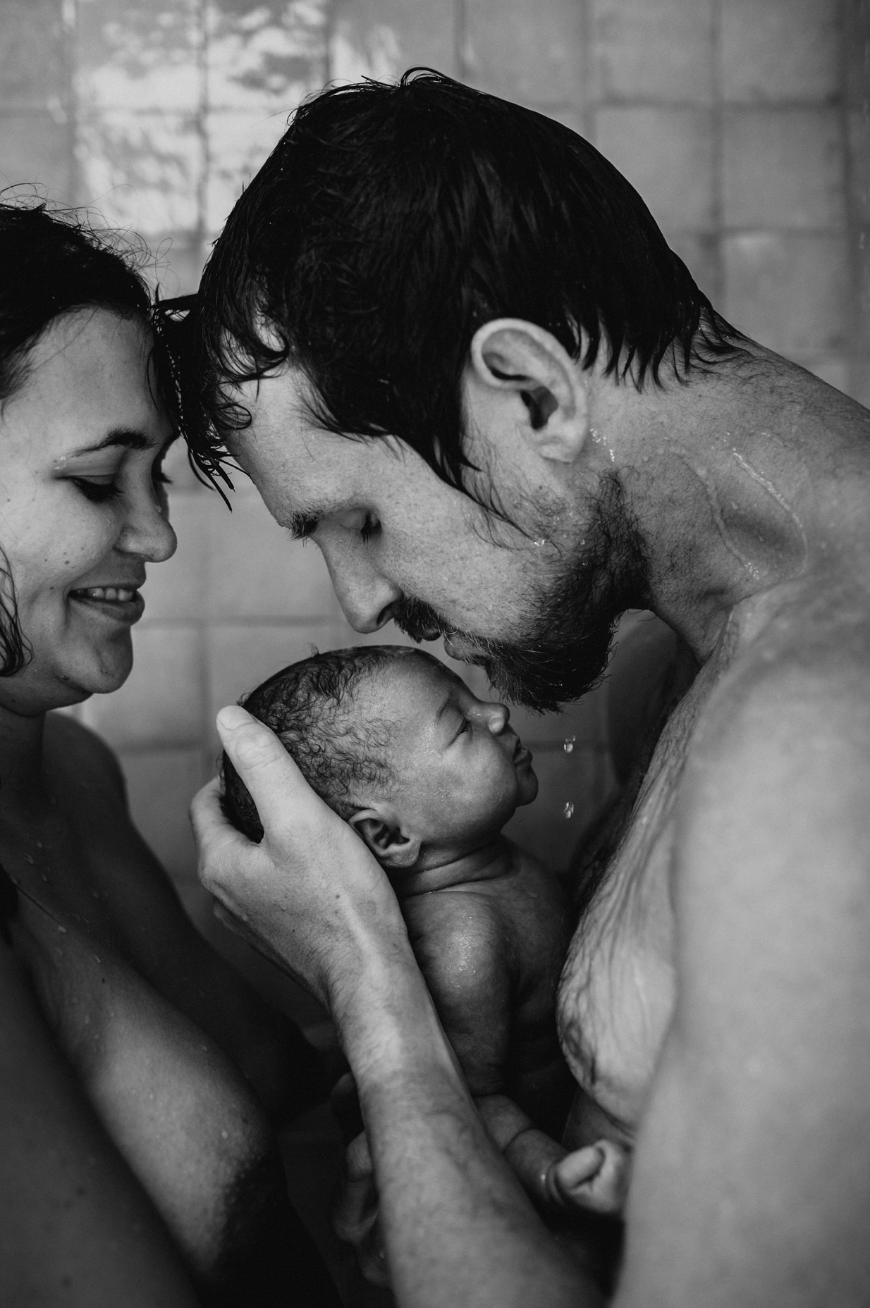 Black and white photo of parents tenderly holding their newborn baby together in a shower, sharing an intimate family moment.