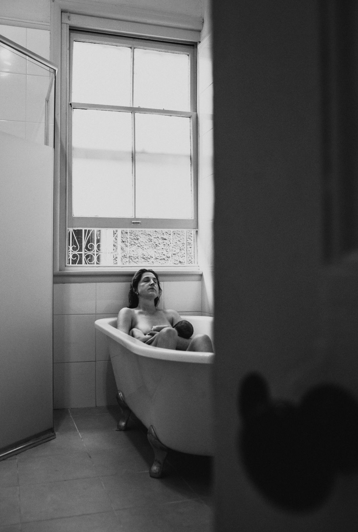 Black and white photo of a woman relaxing in a clawfoot bathtub near a window, holding a newborn baby.