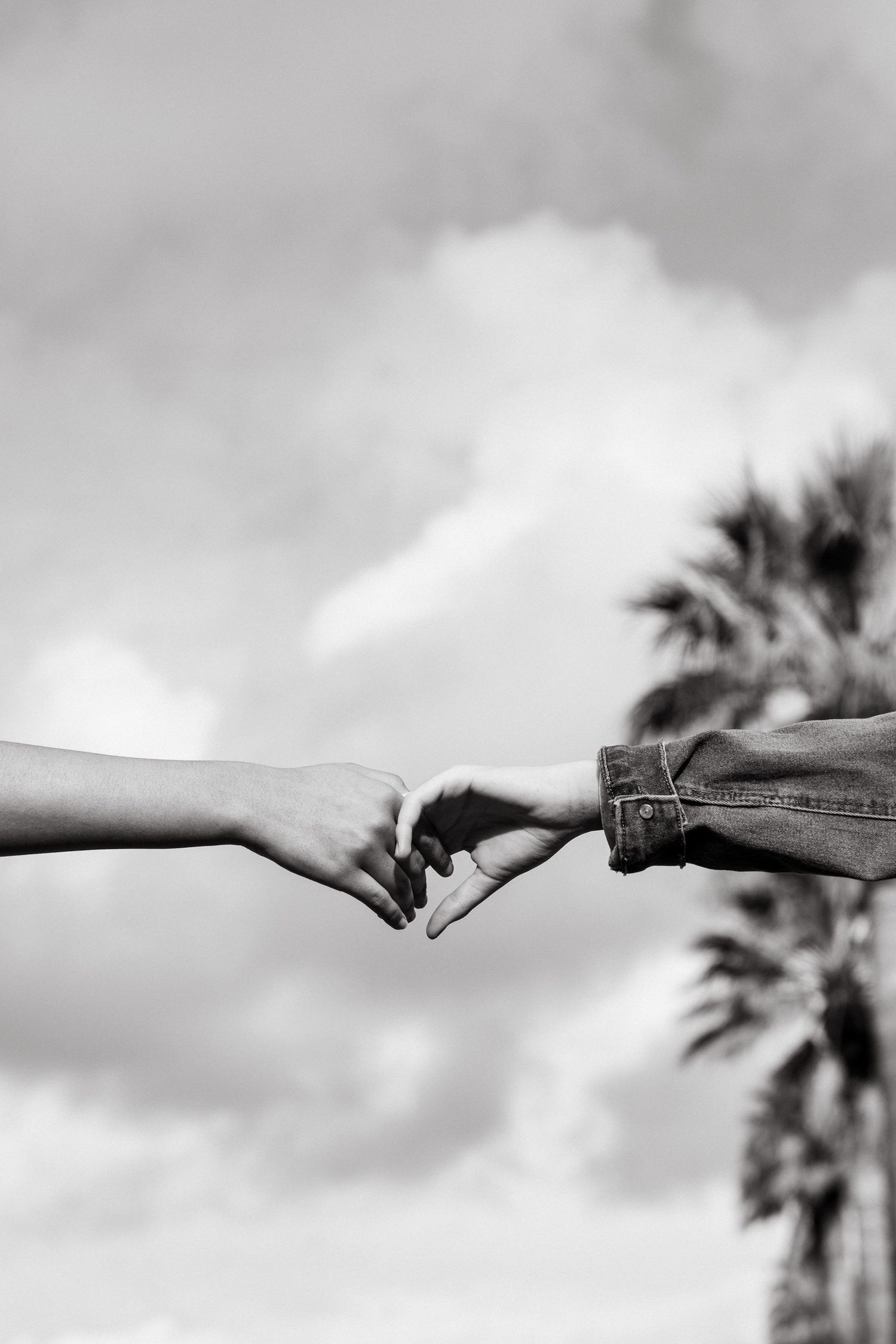 Black and white photo of two hands reaching toward each other with pinky fingers linked against a cloudy sky.