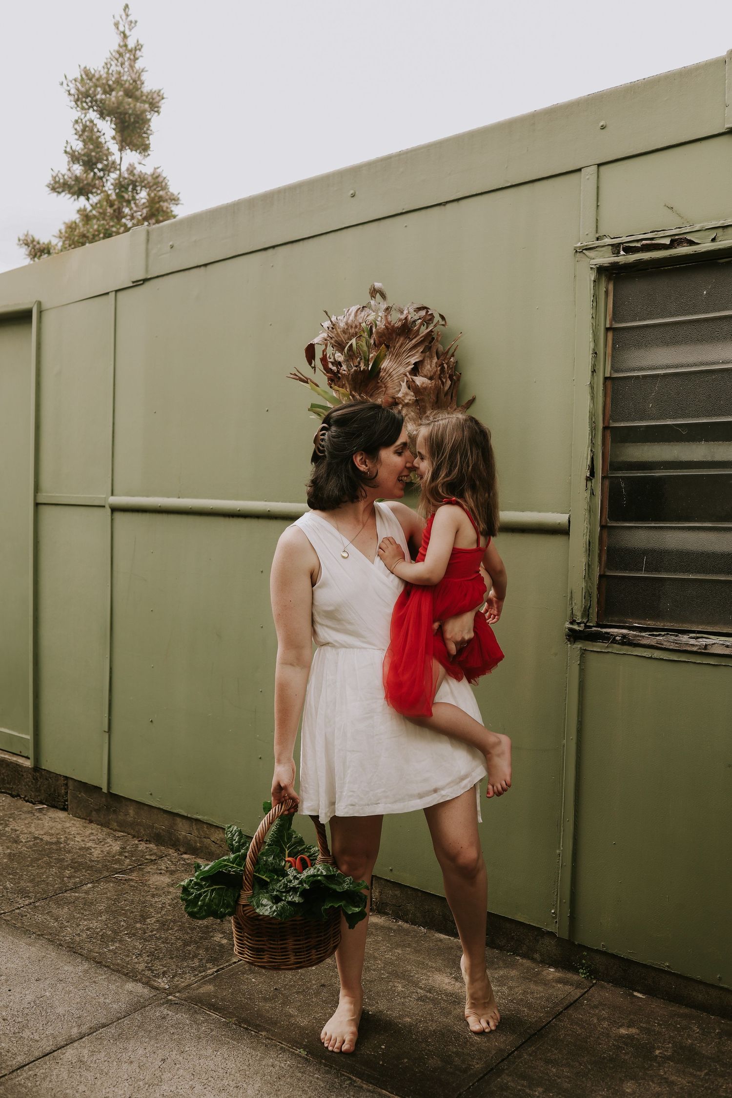 A mother in a white dress holds her young daughter in a red tutu while sharing a kiss outdoors.