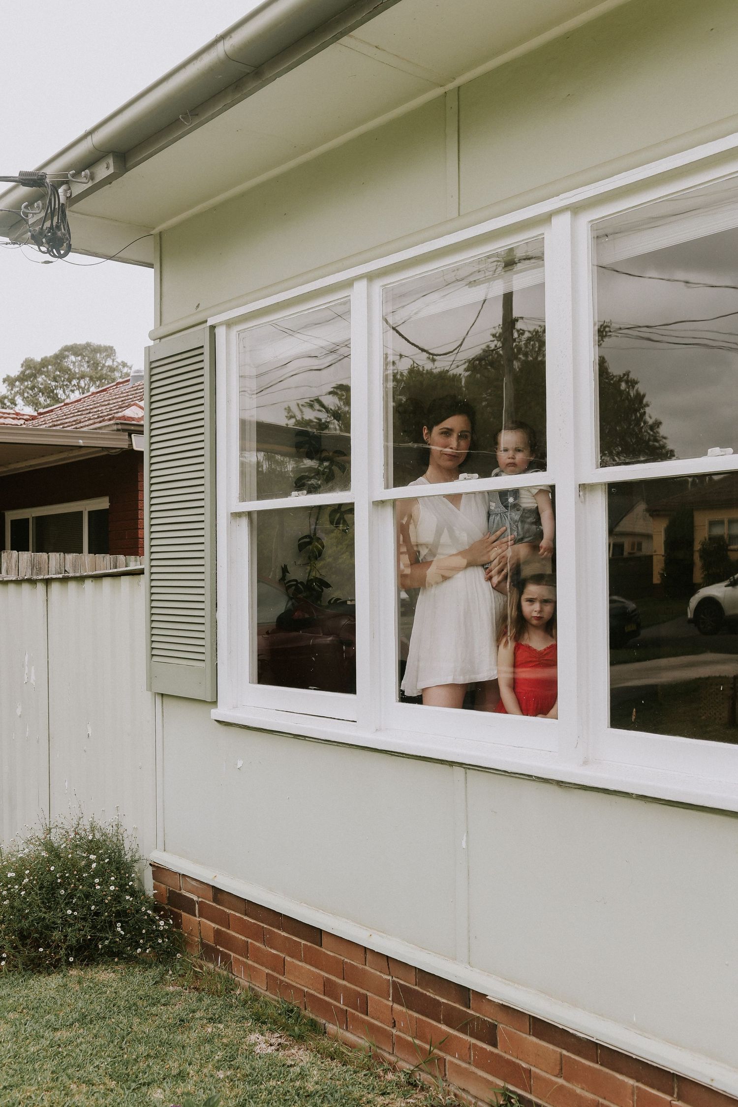 A woman in white dress holds baby while young girl stands beside her, viewed through a house window from outside.
