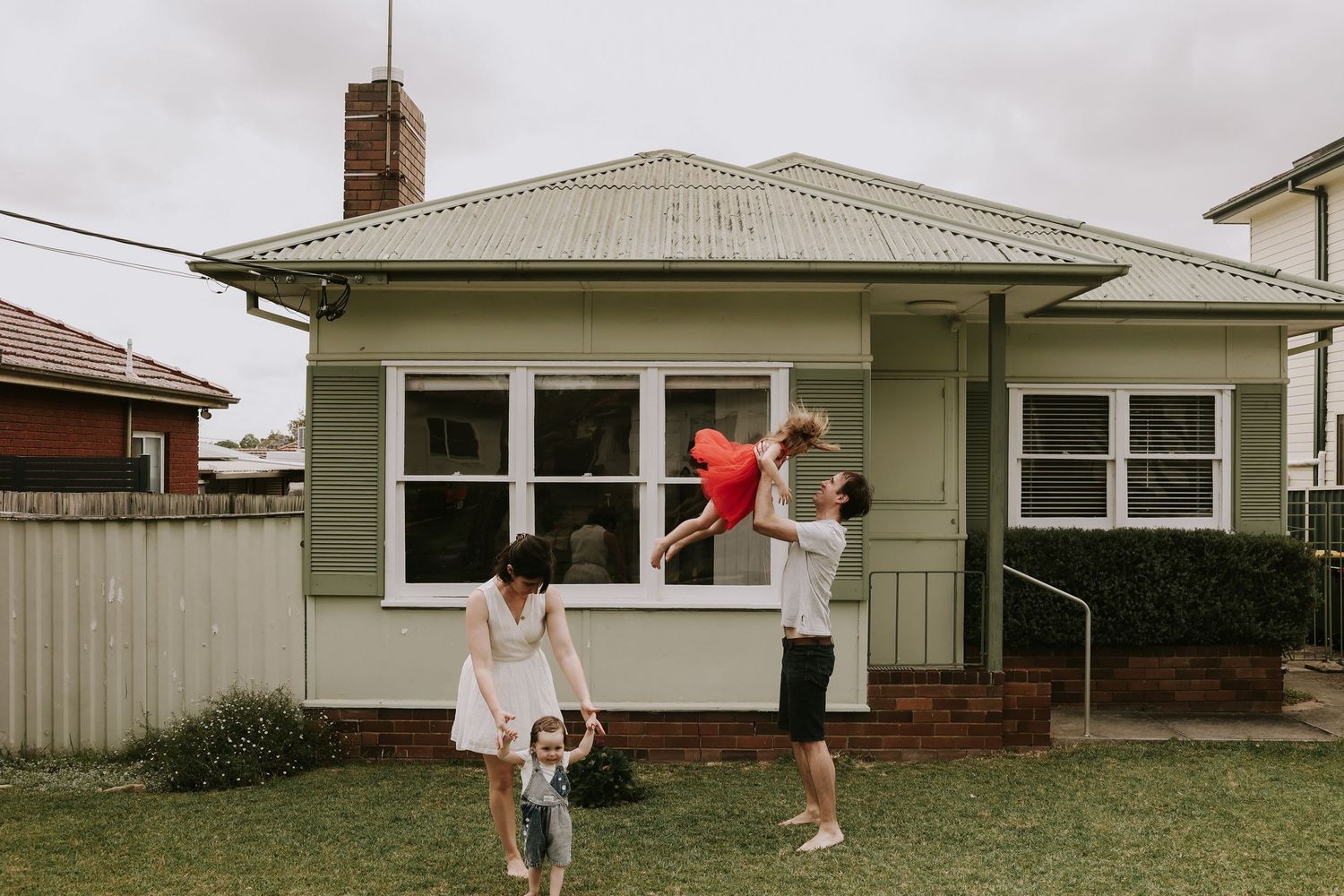 Family playing in front yard of green weatherboard home, parents lifting child in red outdoors.