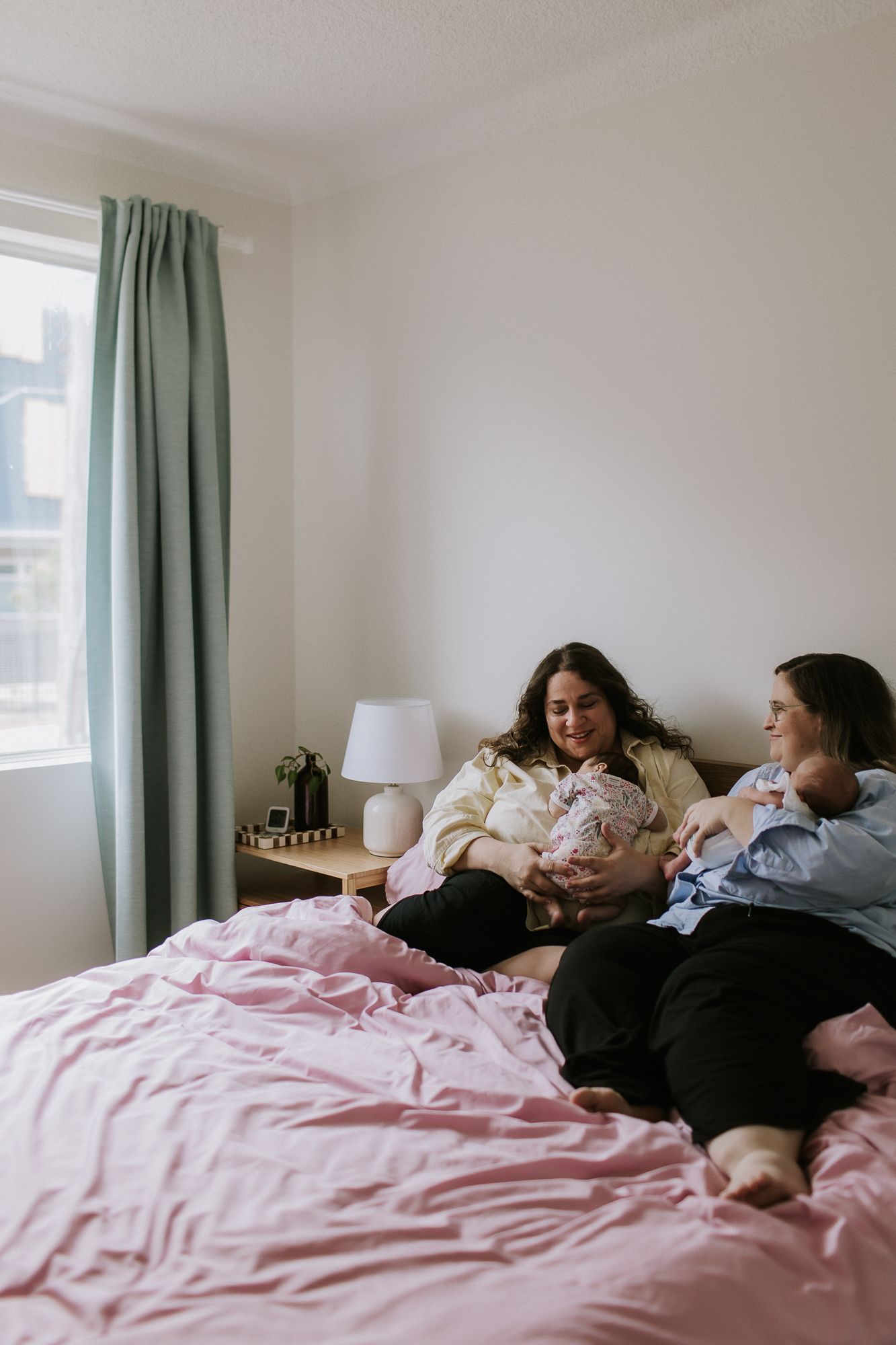 Two women sitting on a pink bed holding newborn babies in a bright bedroom with mint curtains.