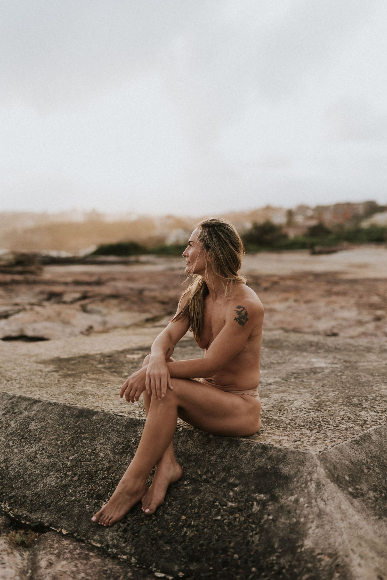 Woman with tattoo sitting on coastal rocks, gazing into distance, with overcast sky and shoreline behind her.